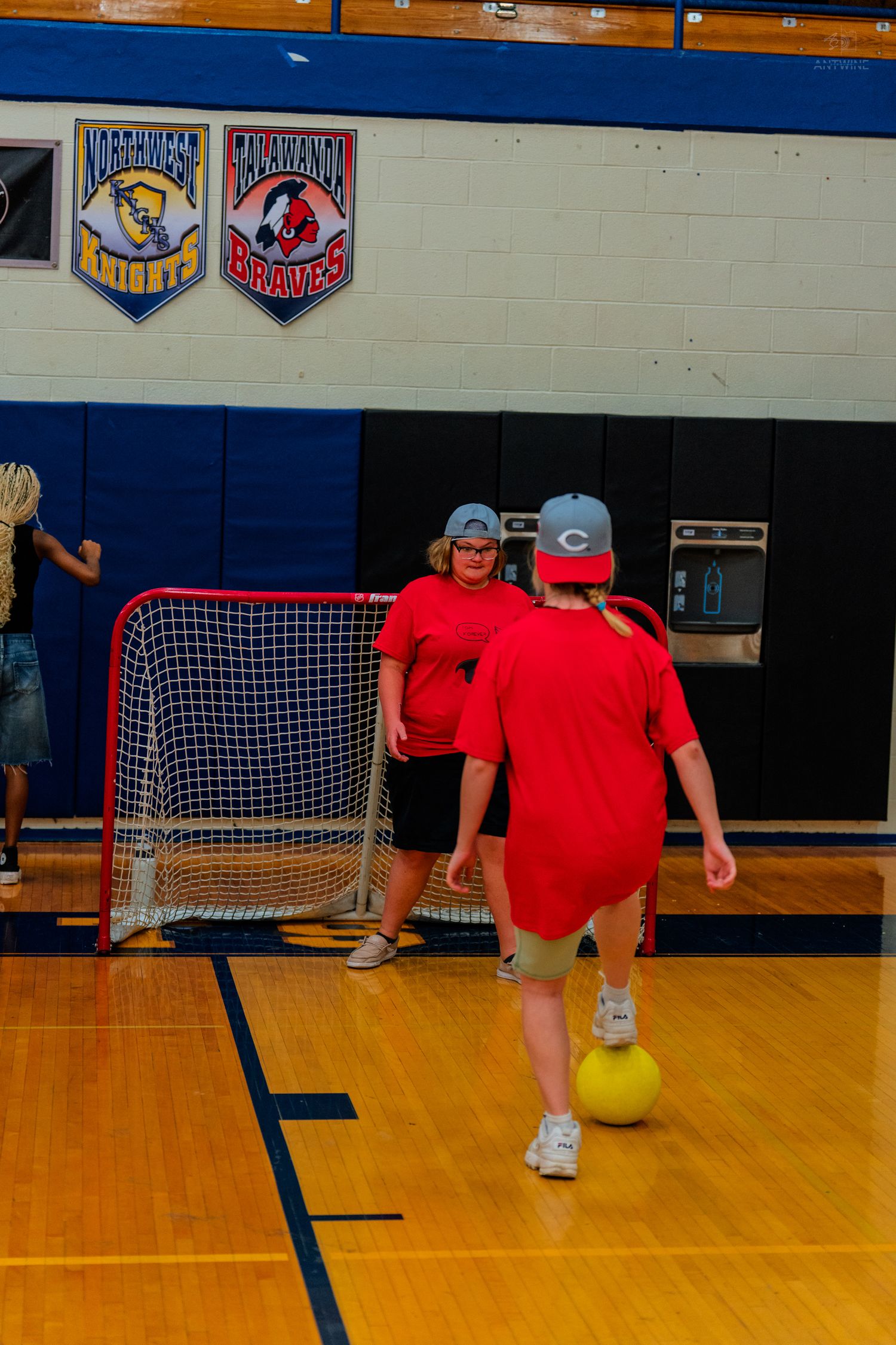 Children in red shirts play indoor soccer in a gymnasium, with a small goal net and yellow ball on hardwood floor.