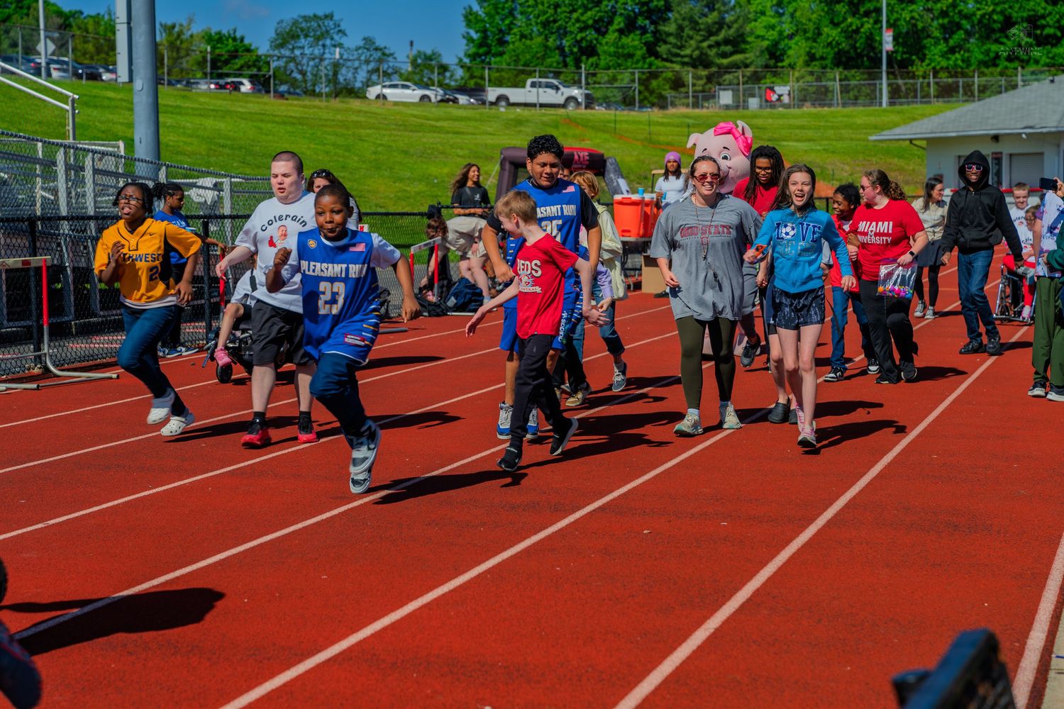 Students running on a red track during an outdoor school field day event with green trees in the background.