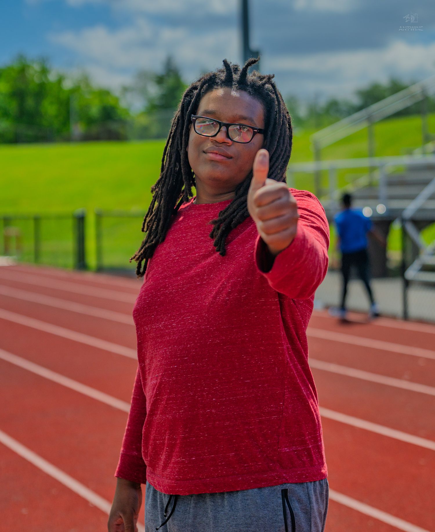 A young Black woman with dreadlocks and glasses gives a thumbs up on a red running track outdoors.