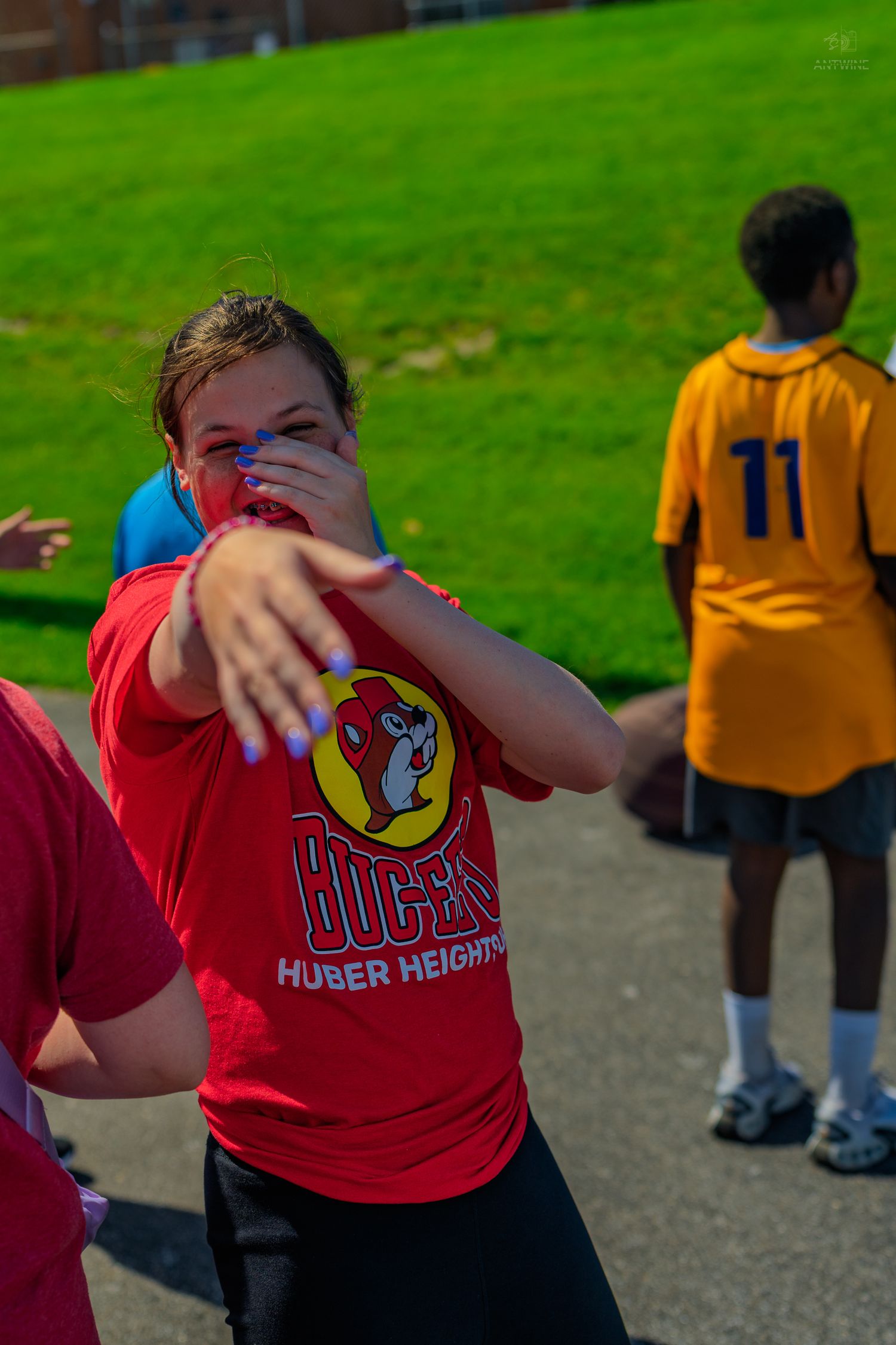 Girl in red Buccees Huber Heights shirt pointing at camera outdoors near green grass with kids playing behind her.