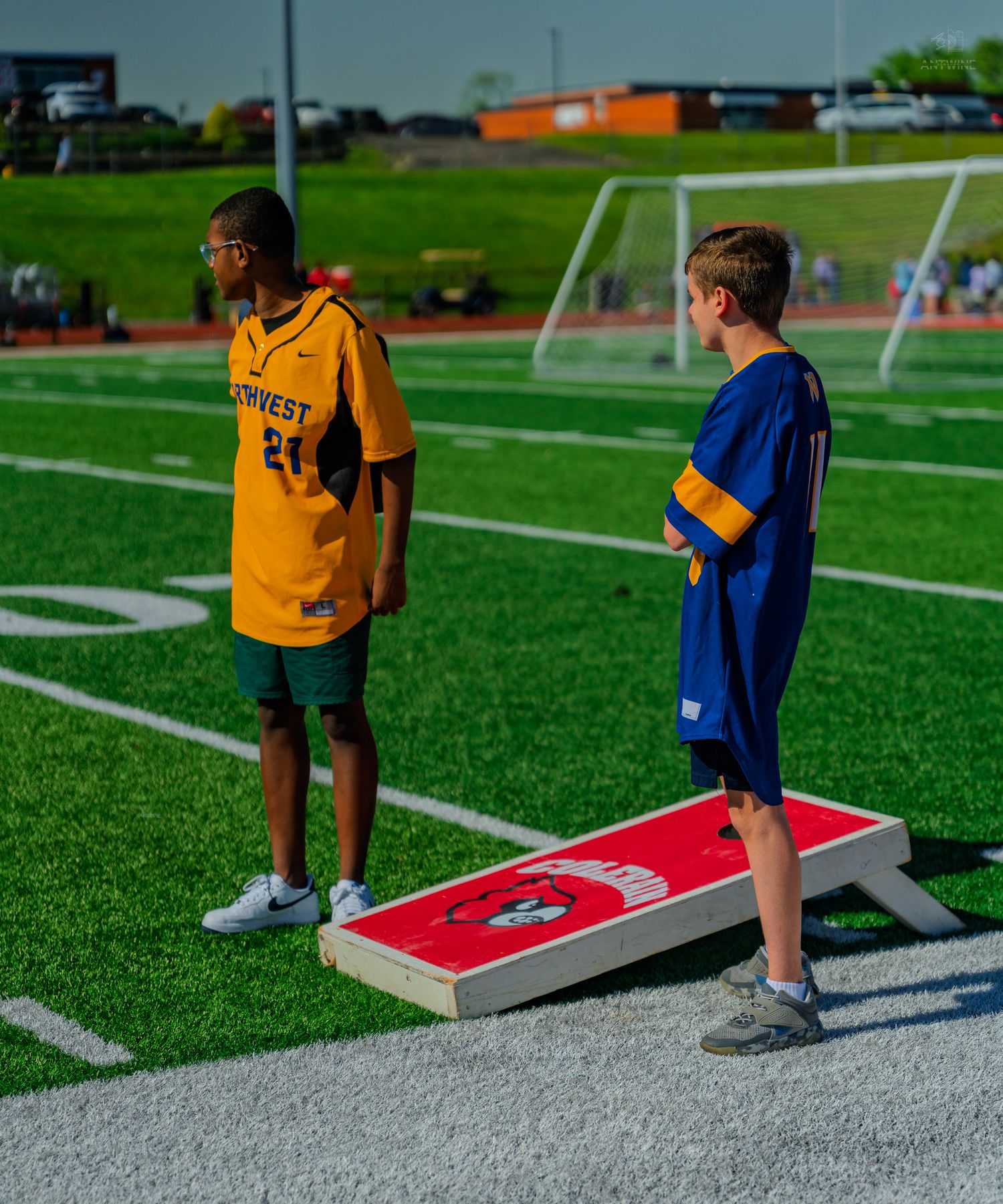 Two boys playing cornhole on a green turf soccer field, one wearing a yellow jersey and one in blue.