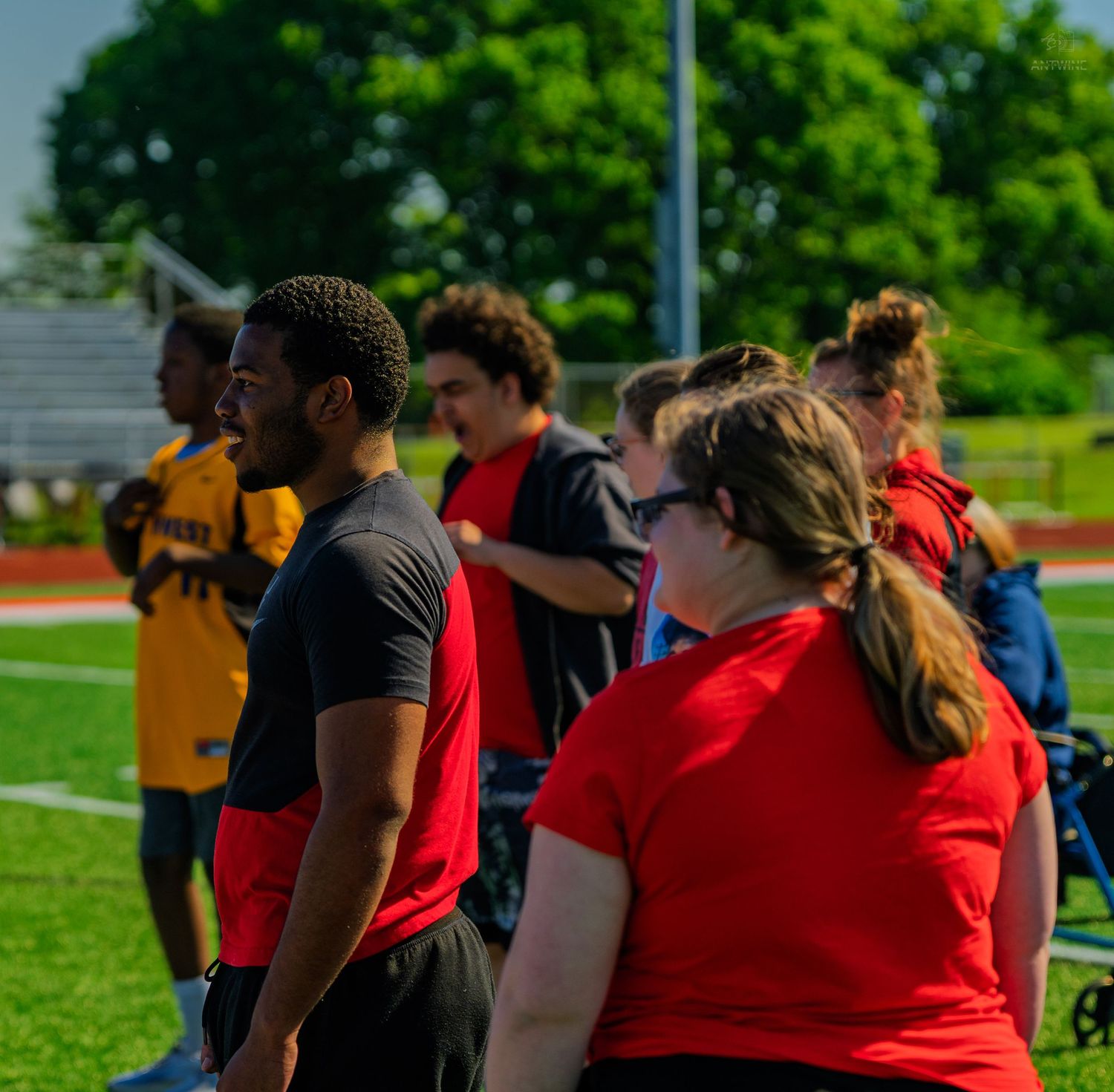 Group of students in red and yellow shirts standing on a track field during an outdoor sports event on a sunny day.