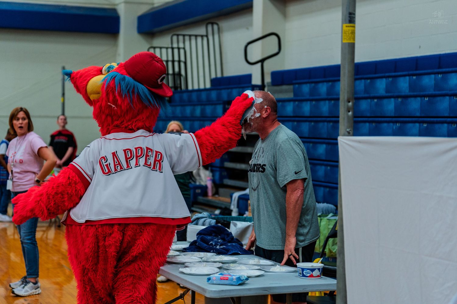 Gapper mascot interacts with a man at an indoor event table in a gymnasium setting.