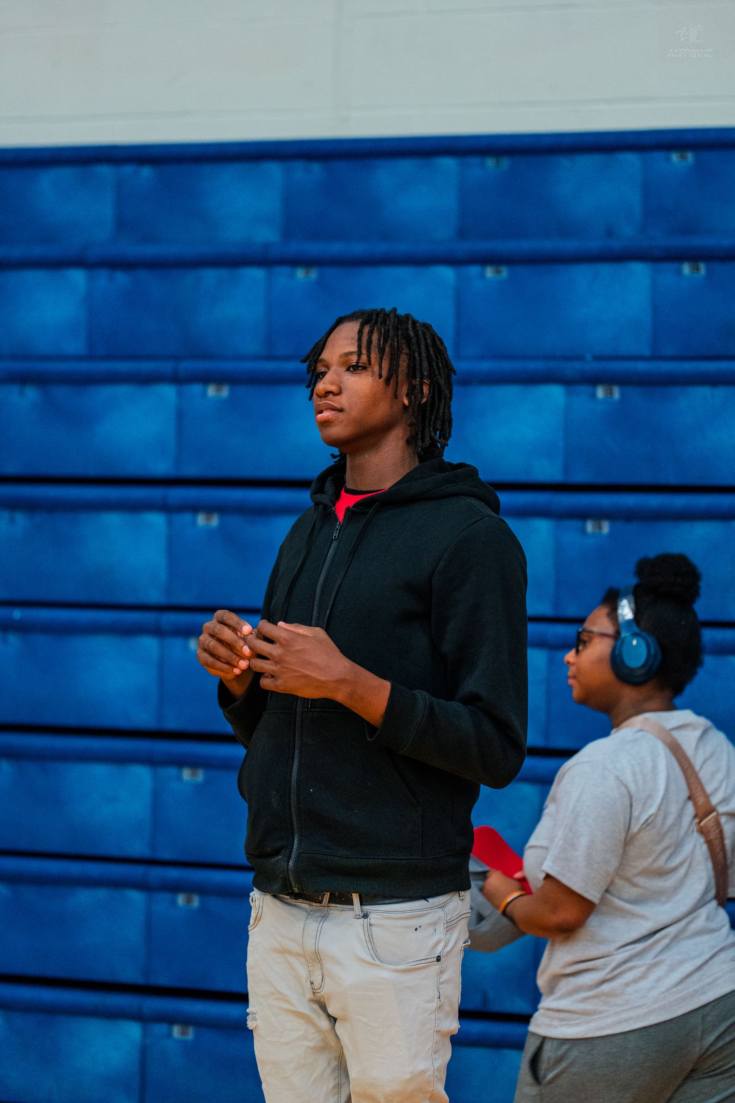 Young man in black hoodie and white pants stands in a gym with blue bleachers, while a woman with headphones stands nearby.
