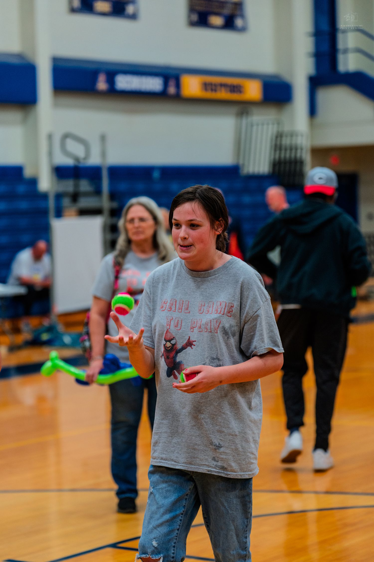 A young person juggling green balls in a school gymnasium while others practice circus skills in the background.
