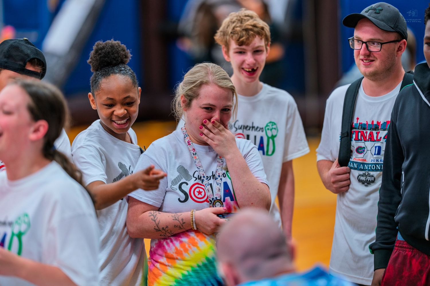 Group of cheerful teens laughing together in a gymnasium, wearing matching white squad t-shirts at a school event.