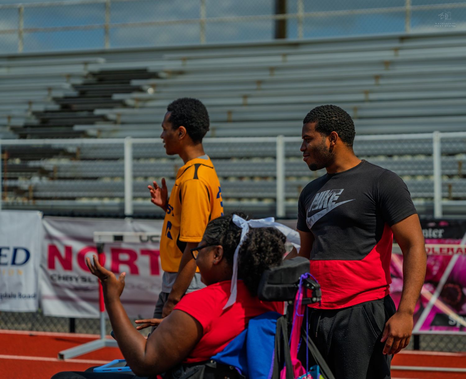 Athletes at a track and field event, including a wheelchair user, gathered near a red and white banner.
