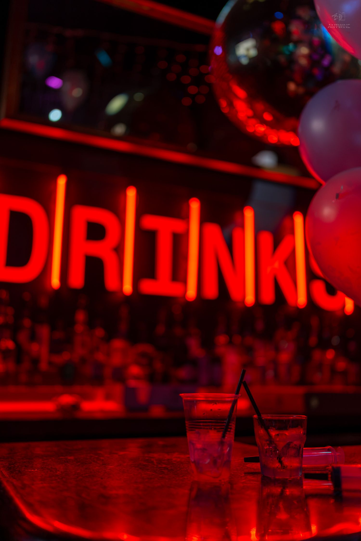 Neon red DRINKS sign glowing behind balloons and glasses on a bar counter in a dimly lit nightclub.