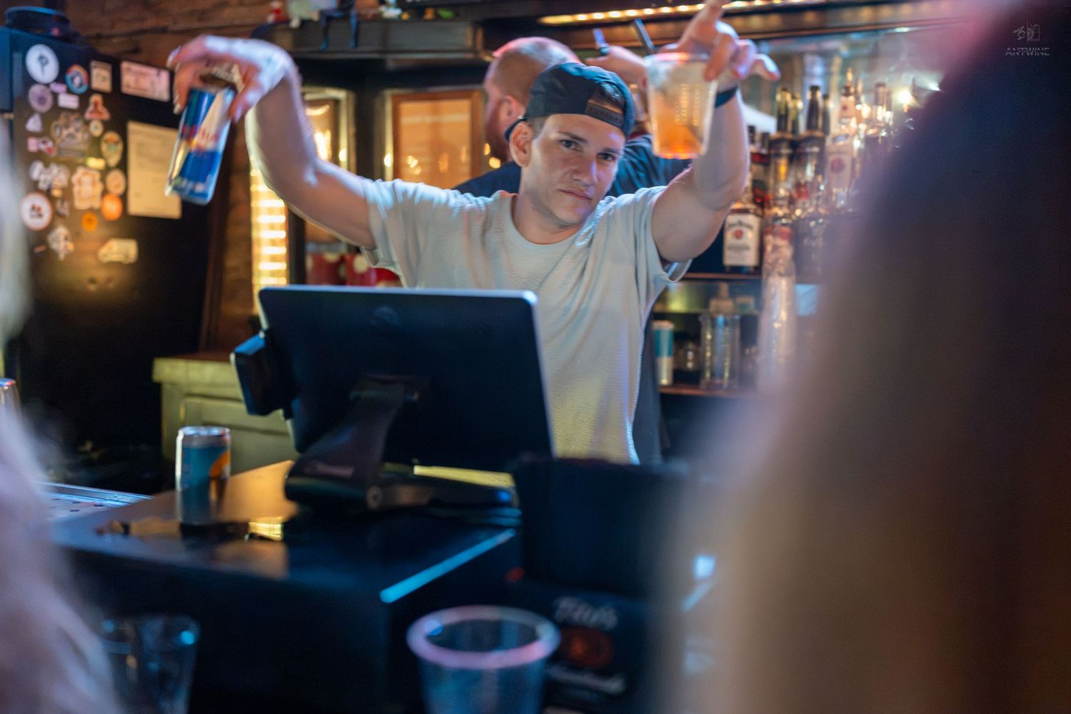 A bartender shakes cocktails behind a bar counter with a POS system, bottles, and dim lighting.