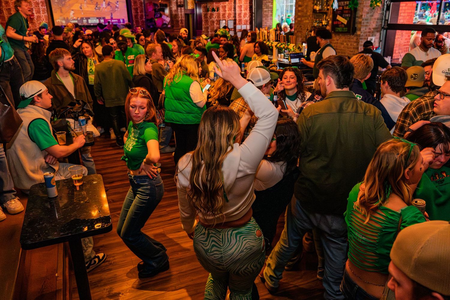 Crowded bar celebrating St. Patrick's Day with people dancing in green outfits on a wooden dance floor.