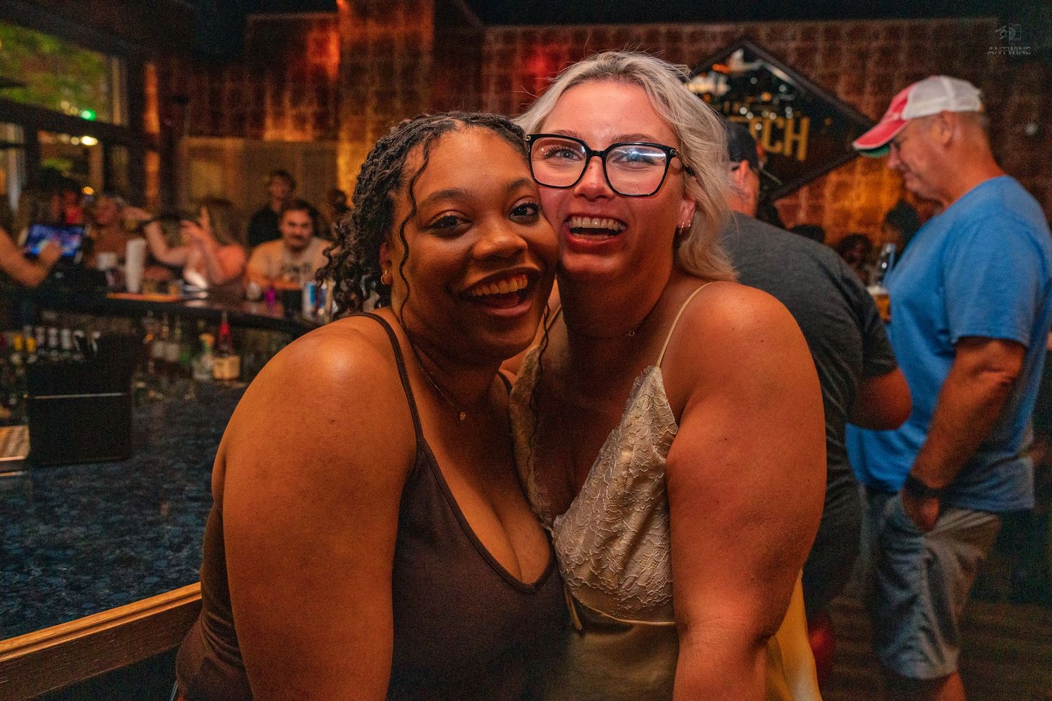 Two women smiling and hugging together in front of a bar with warm lighting.