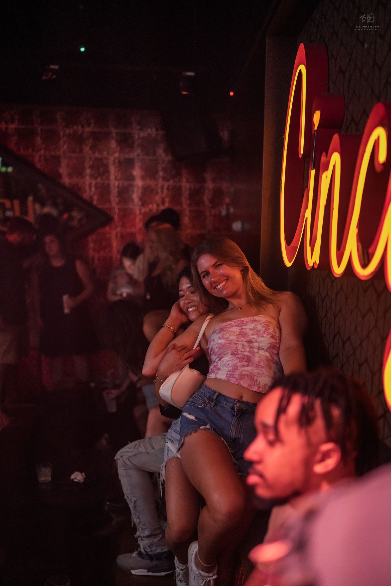 Two women posing and smiling next to a red neon Cincy sign inside a dark nightclub.