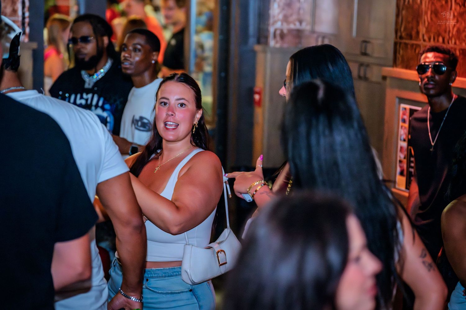 Woman in white crop top chatting with friends in crowded nightlife venue.