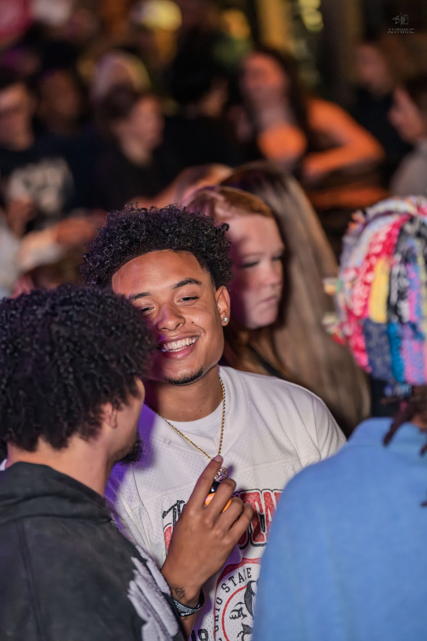 Smiling young man with curly hair in a white graphic tee enjoys a lively nightclub atmosphere with friends.