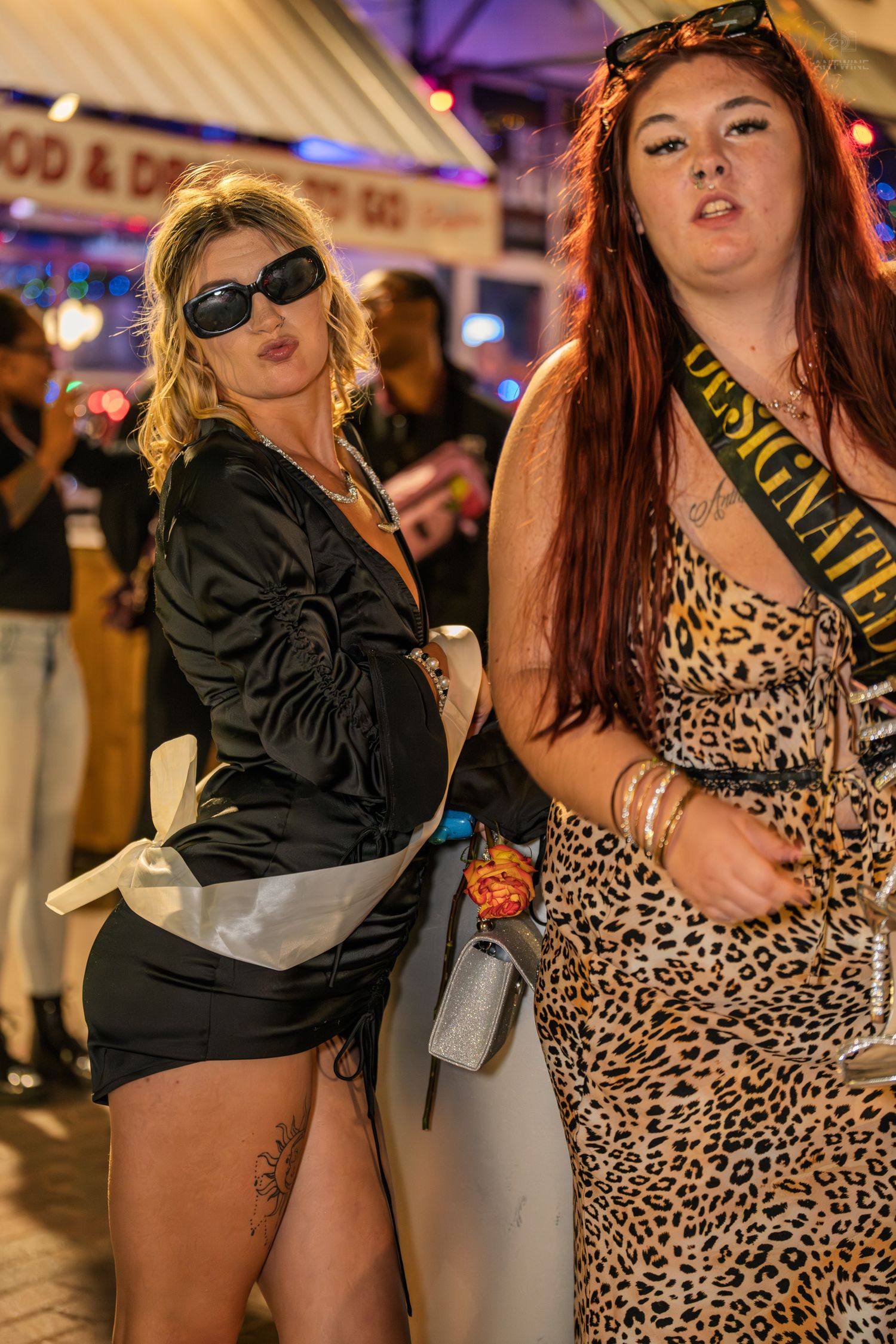Two women pose at night outside a bar, one in black mini dress with sunglasses, other in leopard print dress with sash.