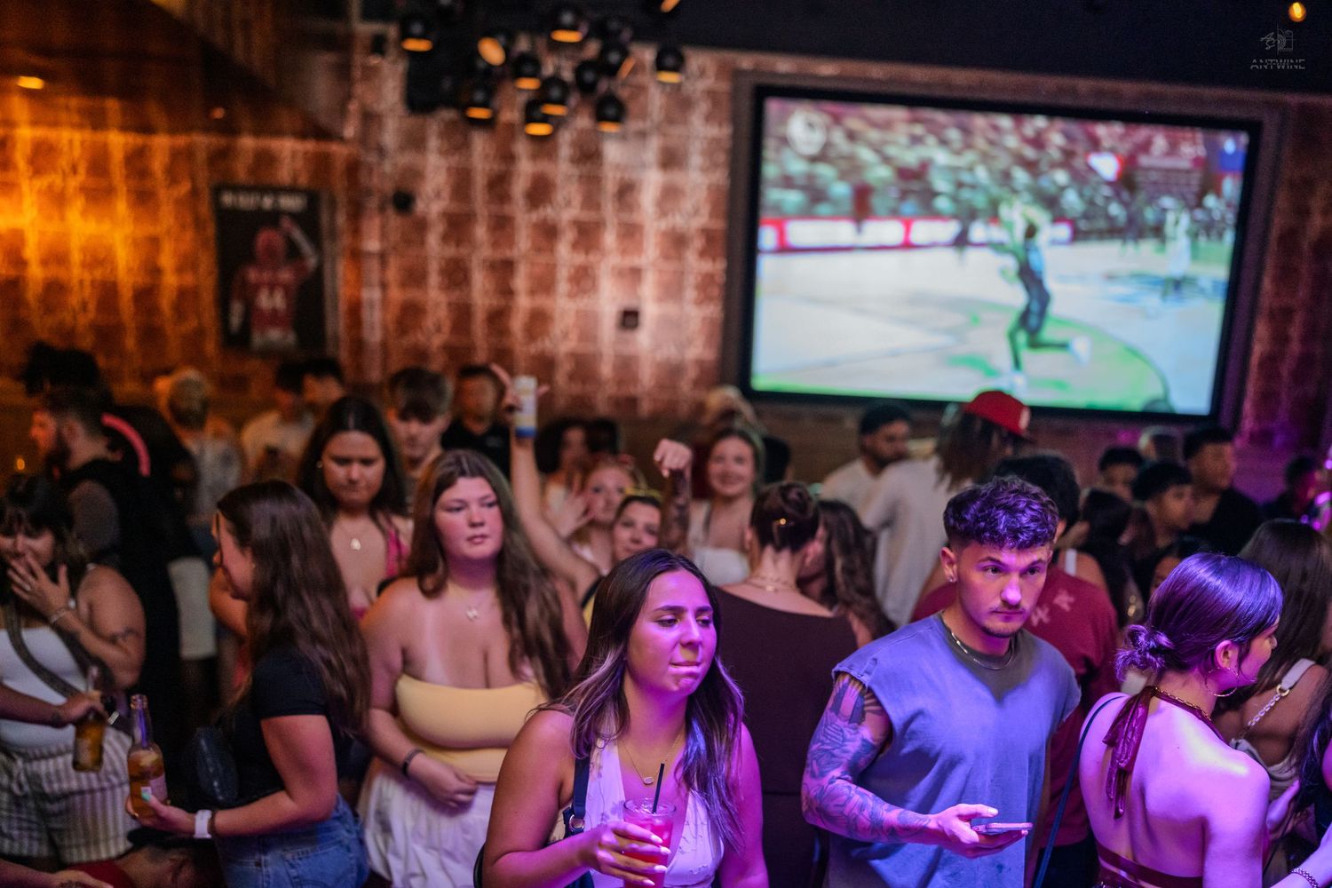 Crowded bar with people dancing and socializing under a large sports game projection screen.