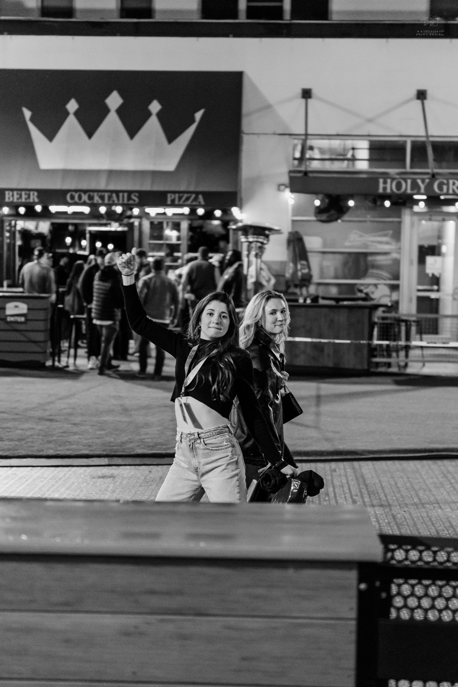 Two women take a selfie outside a bar with a crown logo and Holy Grail sign.