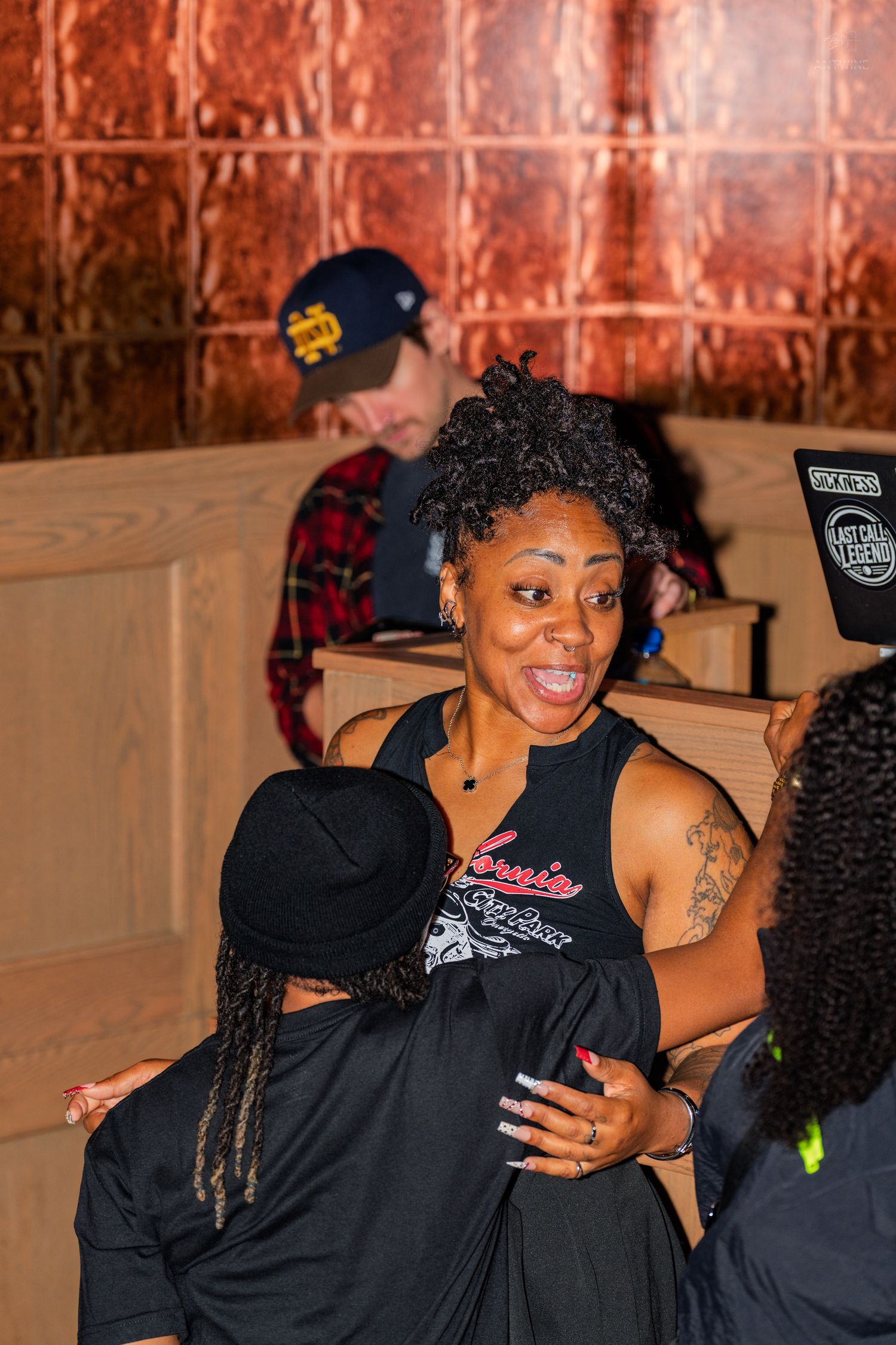 Woman in a sleeveless graphic shirt laughing and hugging friends near a DJ booth at a party.