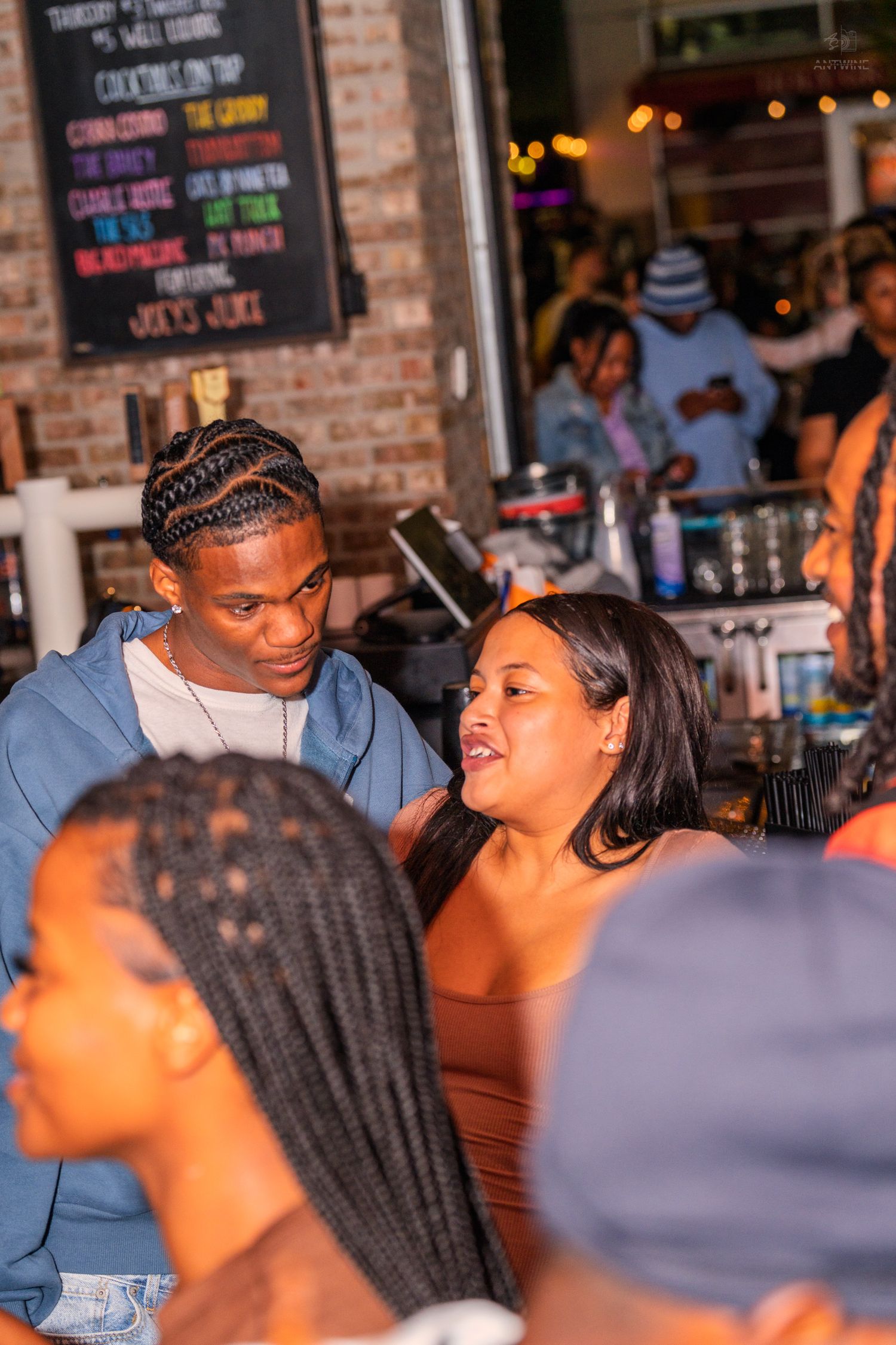 Young couple conversing near a bar with a cocktail menu chalkboard in background.