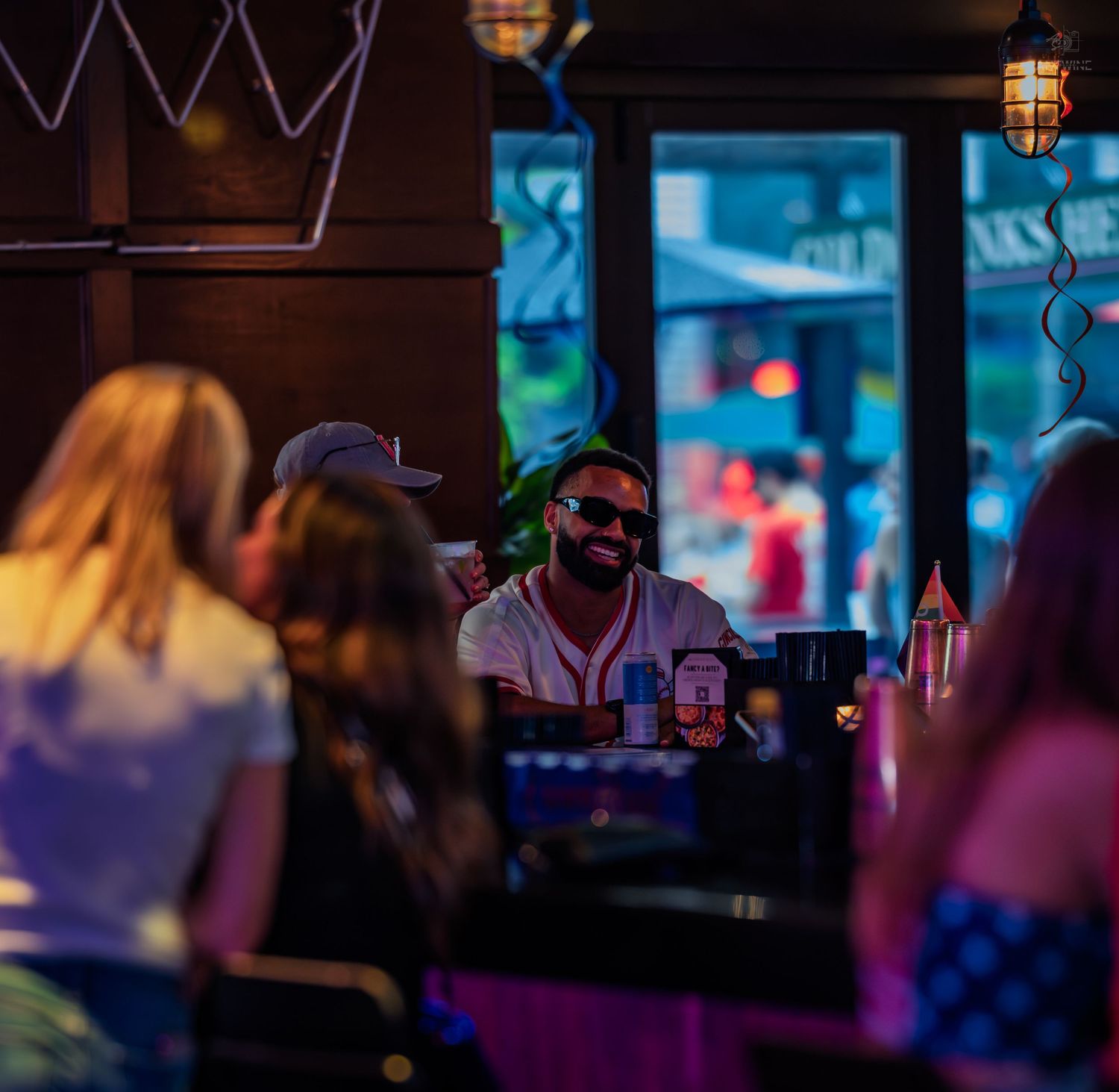 A smiling bearded man in sunglasses enjoys drinks with friends at a warmly lit bar.