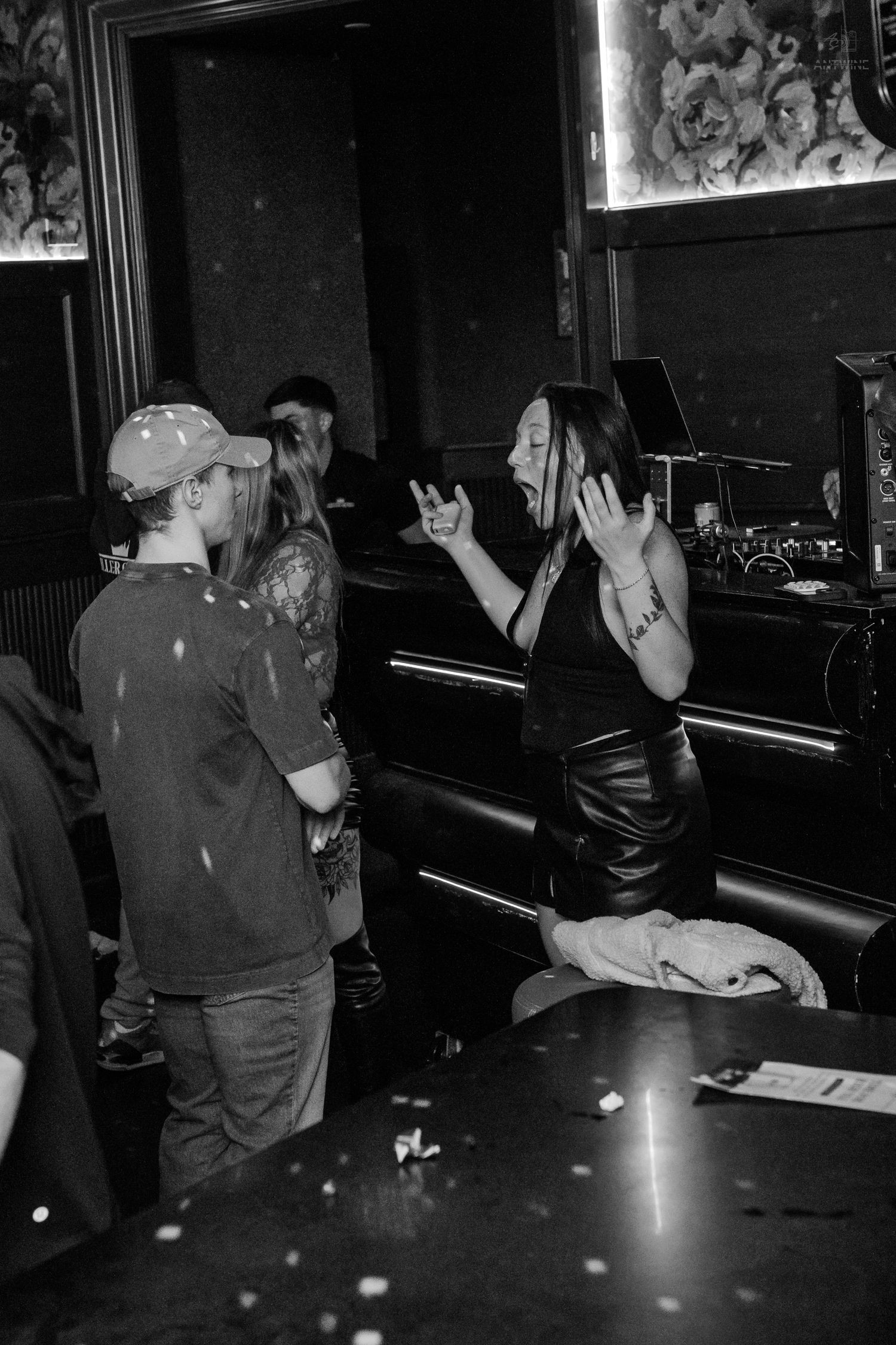 A woman in a leather skirt makes an expressive gesture at a dark nightclub bar while talking to others.
