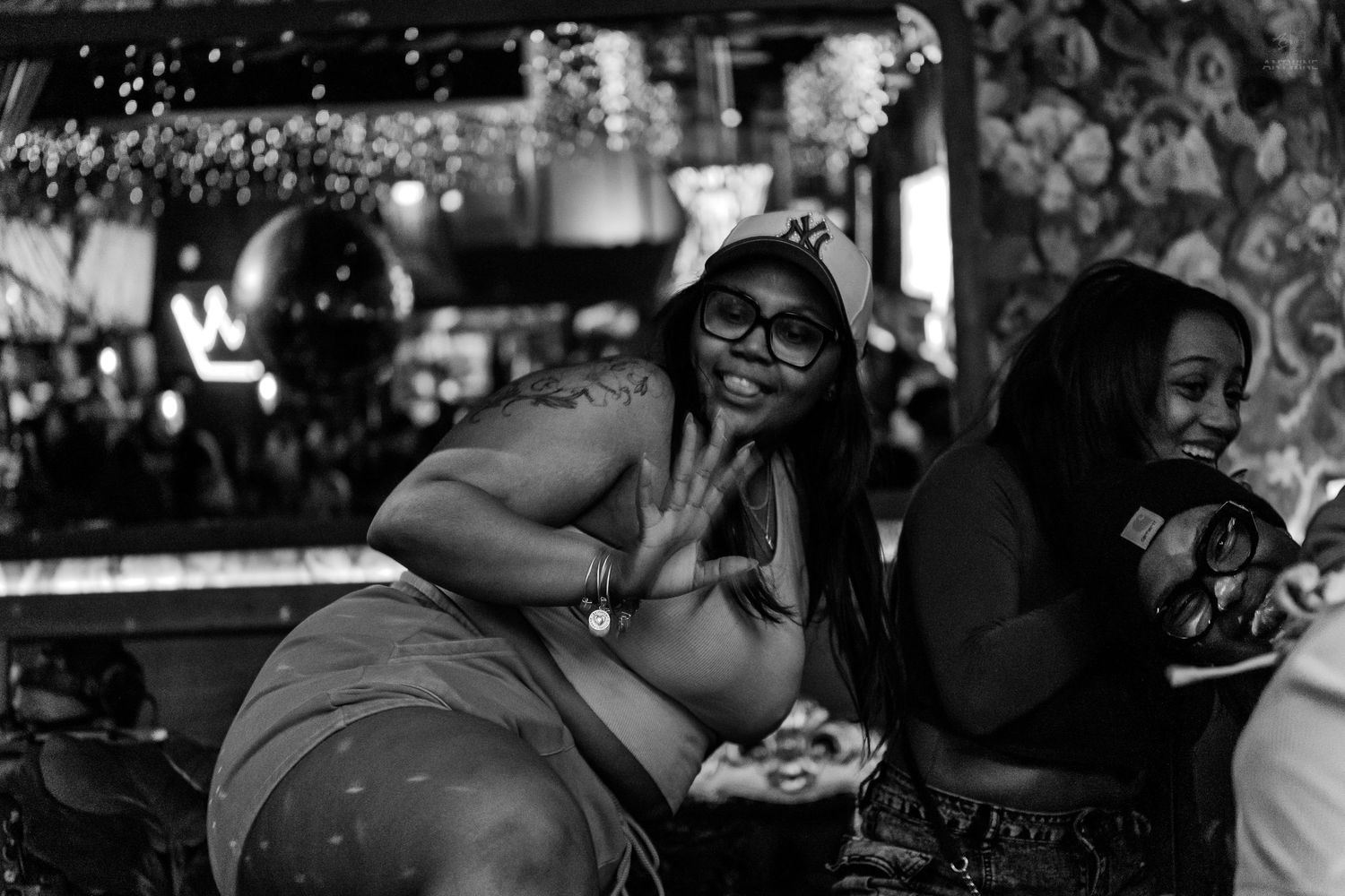 Two women laughing and posing at a lively bar, one wearing glasses flashing a peace sign in black and white photo.