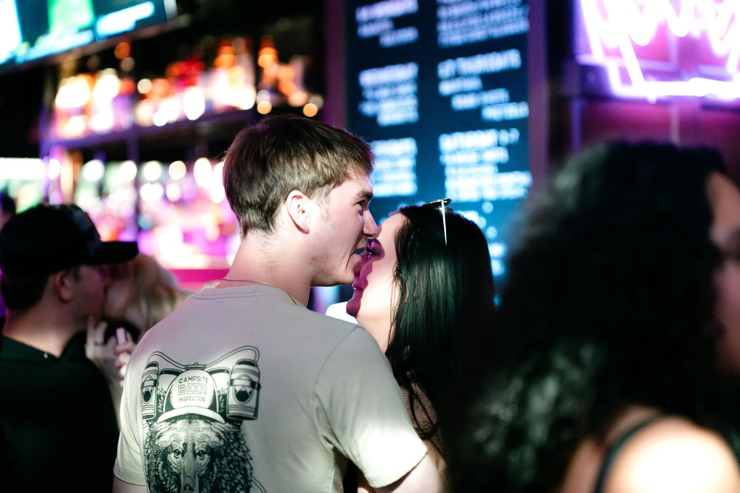 Couple sharing a kiss at a busy nightclub with colorful neon signs in the background.