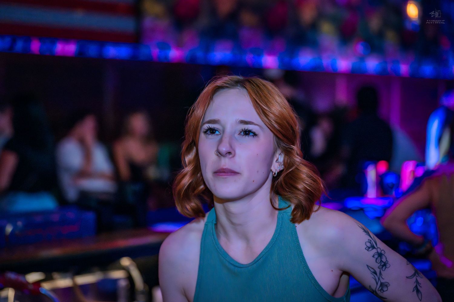 Young redhead woman with tattoo gazes upward under neon lights at a nightclub.