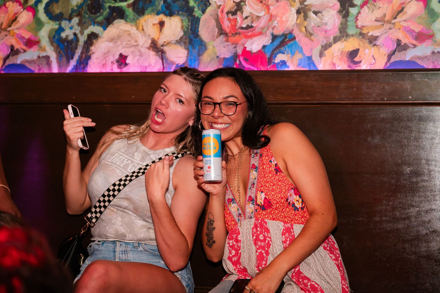 Two women laughing and posing together at a vibrant bar with floral wall art.