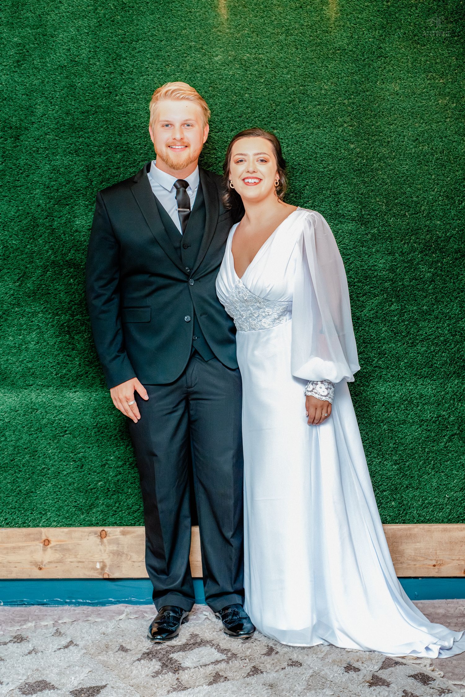 Bride in white gown and groom in black suit pose together in front of a green hedge wall backdrop.