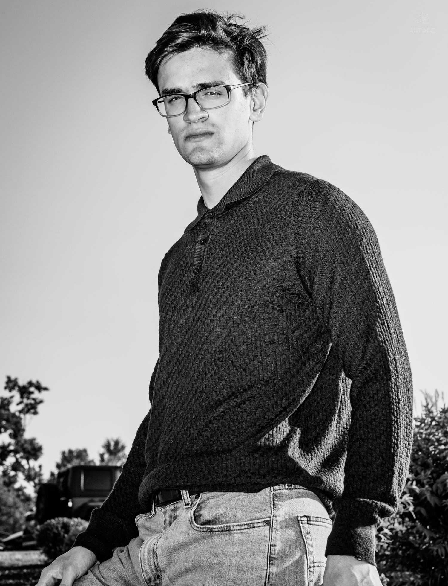 Black and white portrait of a young man wearing glasses and a textured knit sweater with jeans outdoors.