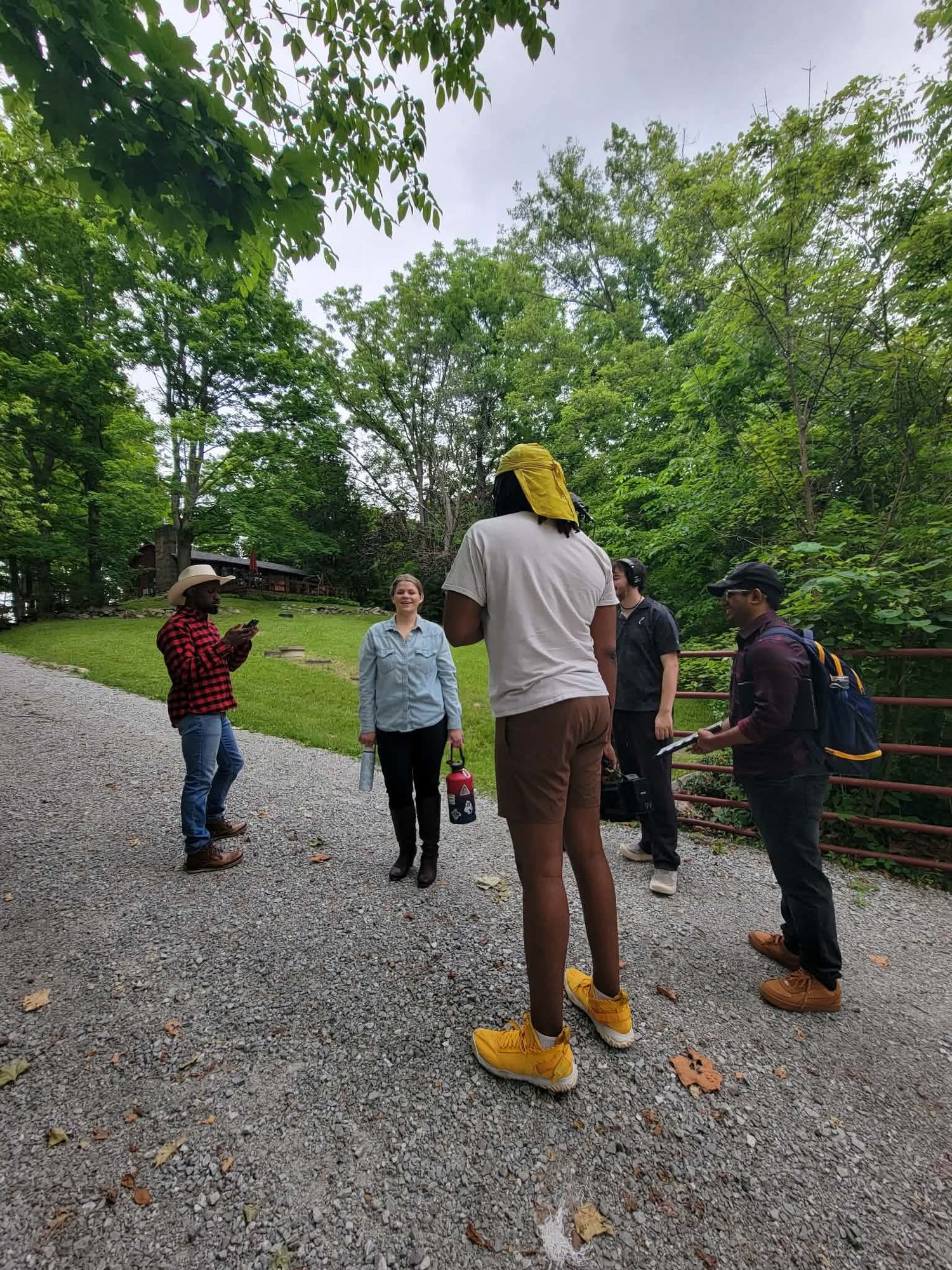 A film crew discussing a scene outdoors on a gravel path surrounded by lush green trees.