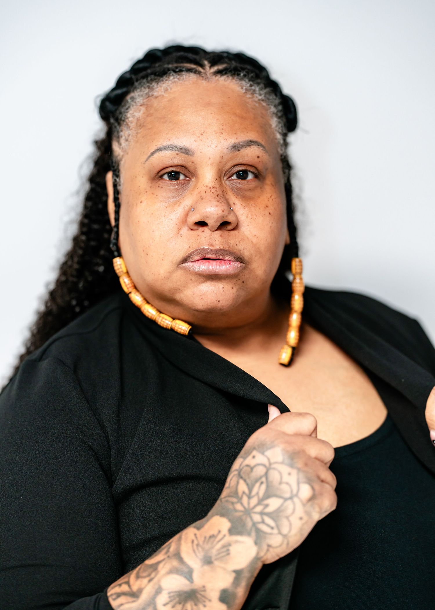 Black woman with braided hair and yellow hoop earrings showing floral arm tattoo, serious expression.