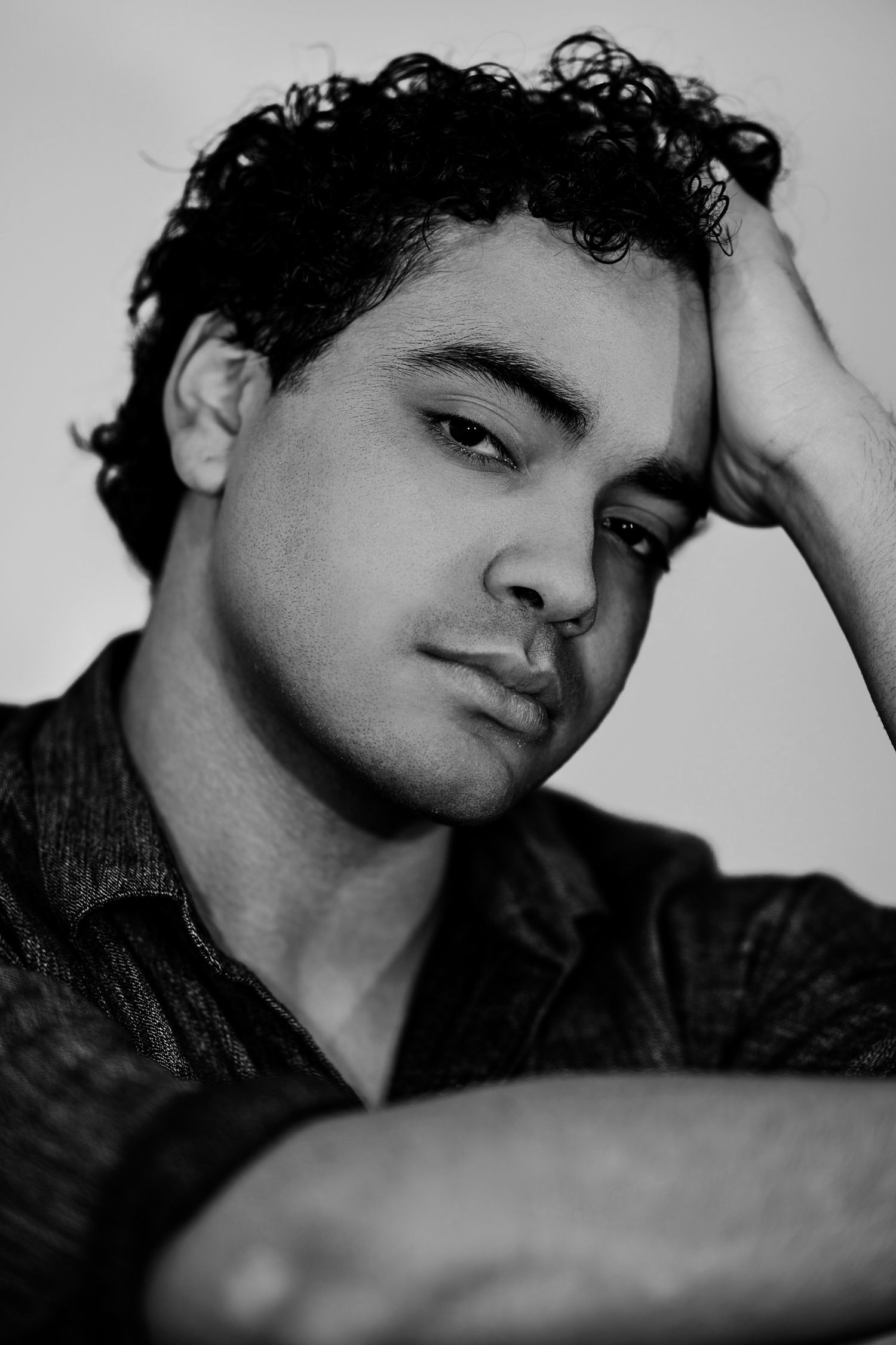 Black and white portrait of young man with curly hair resting hand on head.