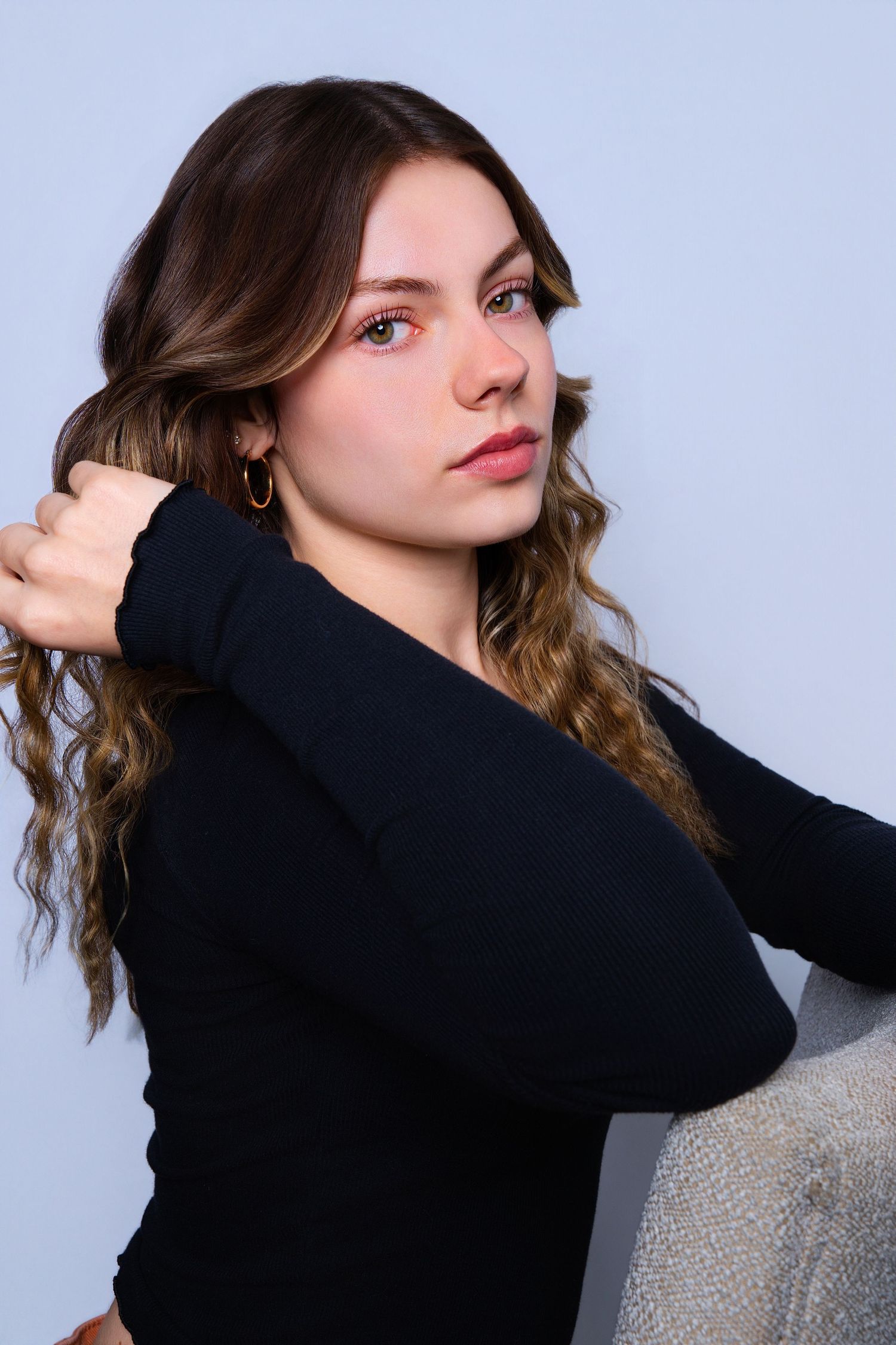 Young woman with wavy hair in black top adjusts hair while posing against gray background.
