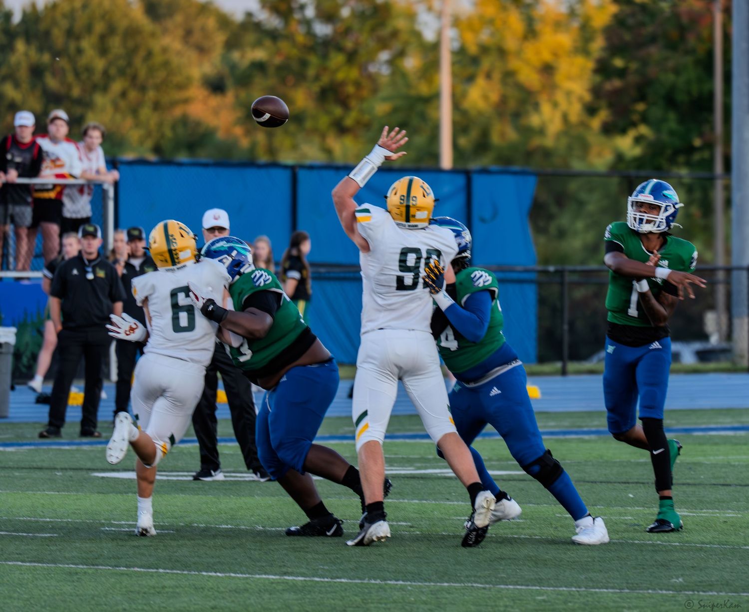 A quarterback in gold uniform throws a pass under pressure during a night football game.
