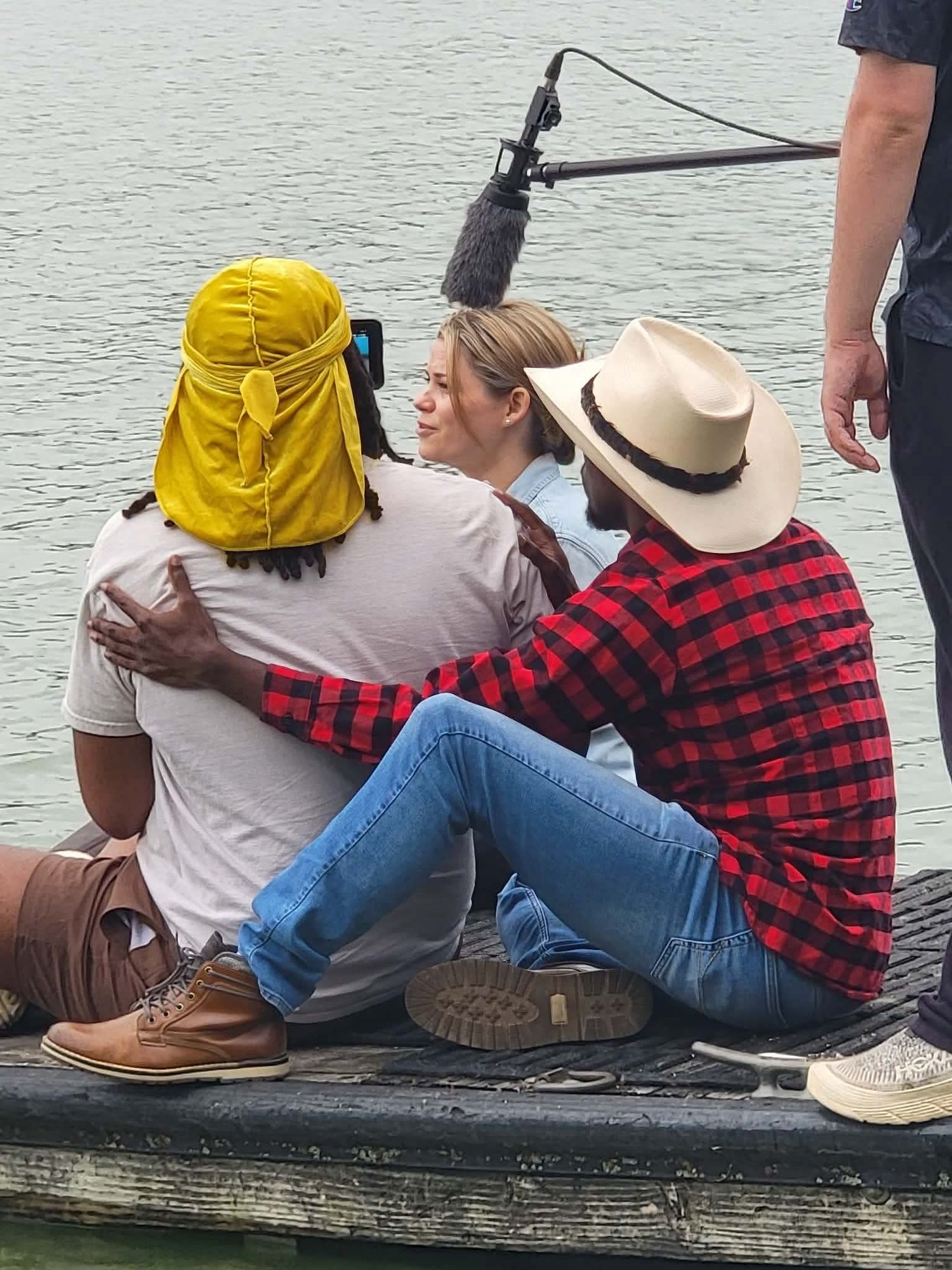 Three people sitting on a dock by the water, with a boom microphone overhead during a film shoot.