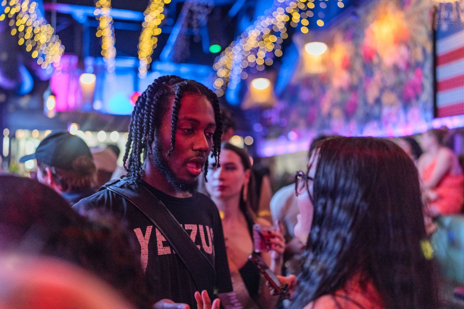 Man in Yeezus shirt socializing at a colorful nightclub with festive lighting.