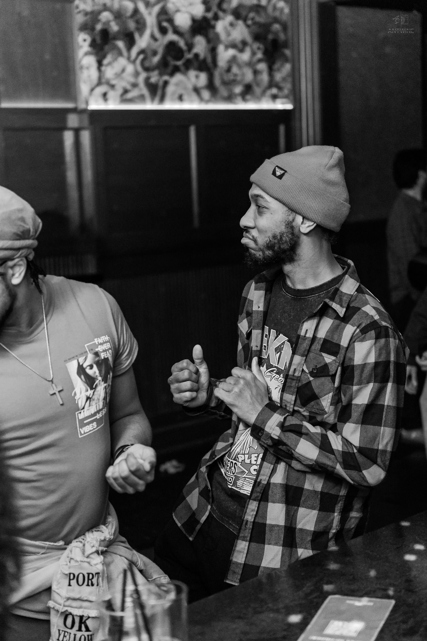 Two men socializing at a bar, one wearing a plaid shirt and beanie hat in black and white photography.