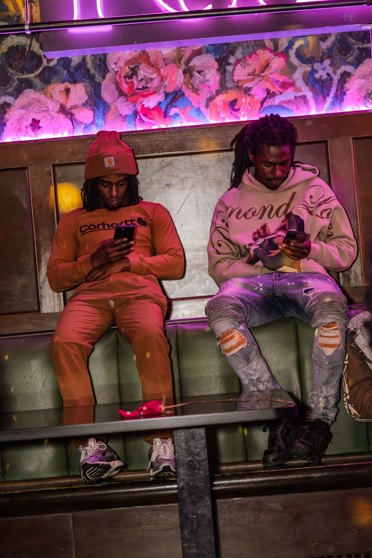 Two men in streetwear sit on green booth seating scrolling phones under pink neon club lighting.