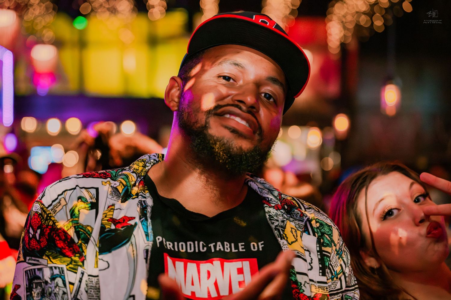 Smiling man wearing a Marvel t-shirt and snapback hat at a vibrant nightclub.