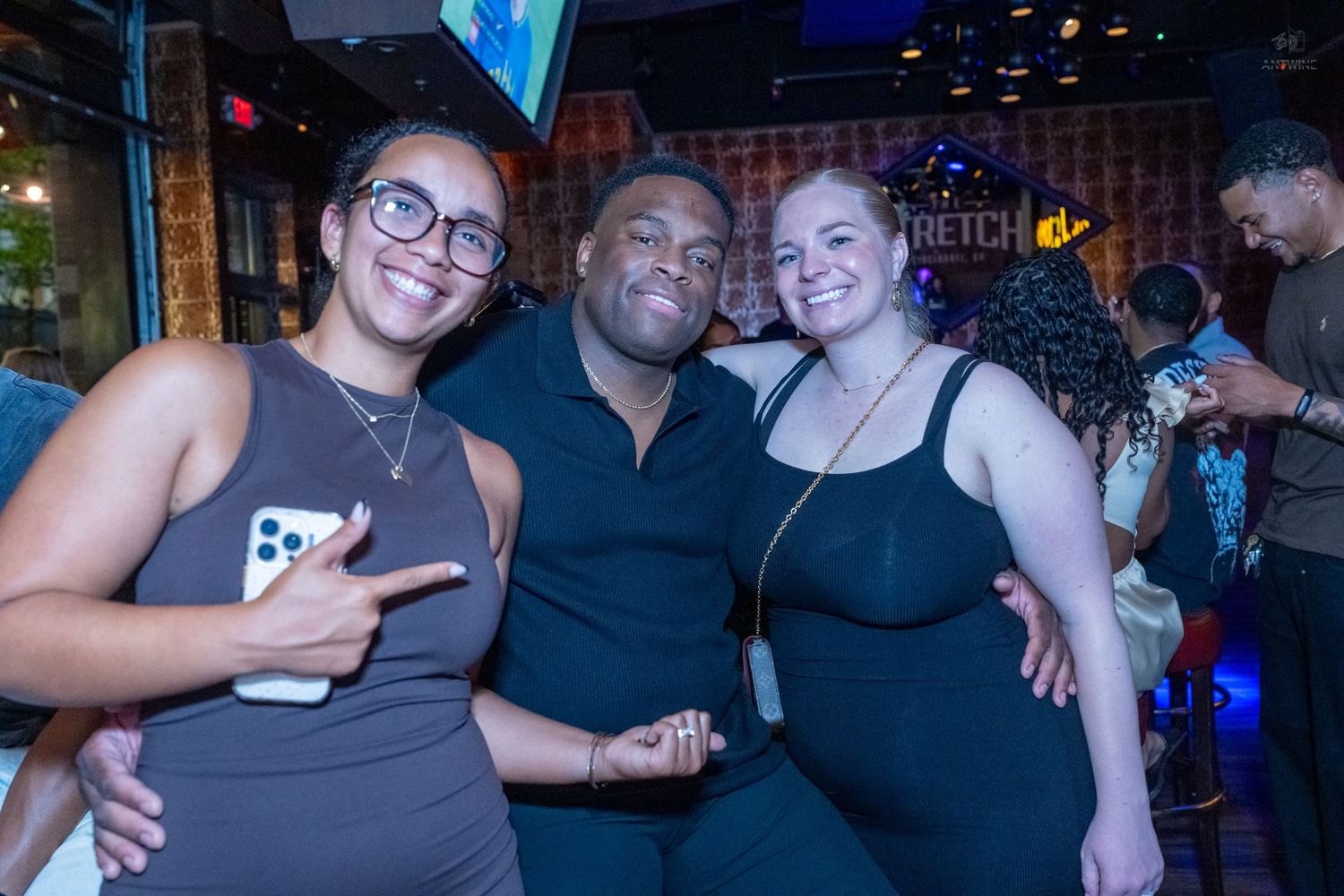 Three friends pose together smiling at a bar, with a Stretch sign visible in the background.