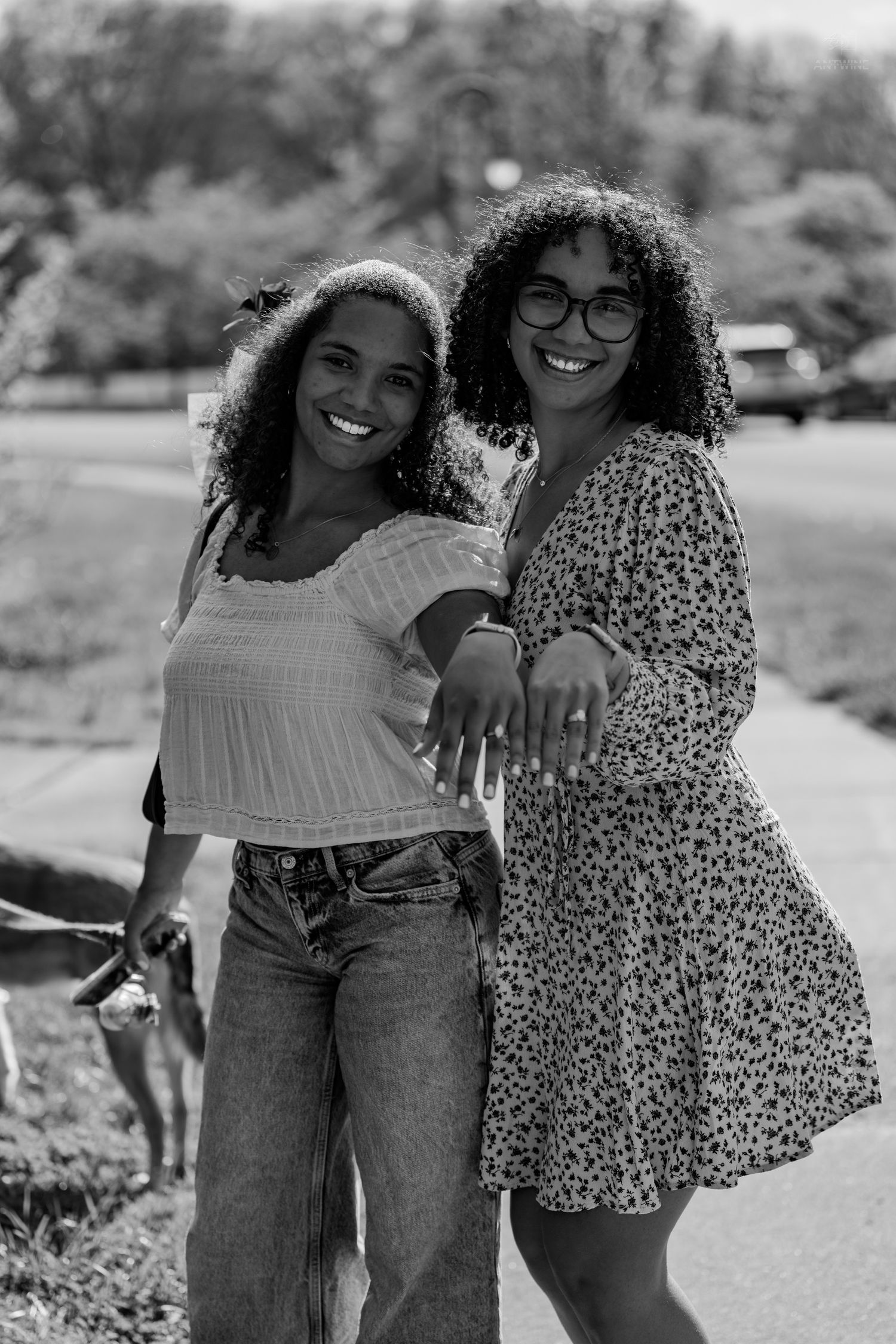 Two smiling women in black and white photo showing off engagement rings while standing in a park.