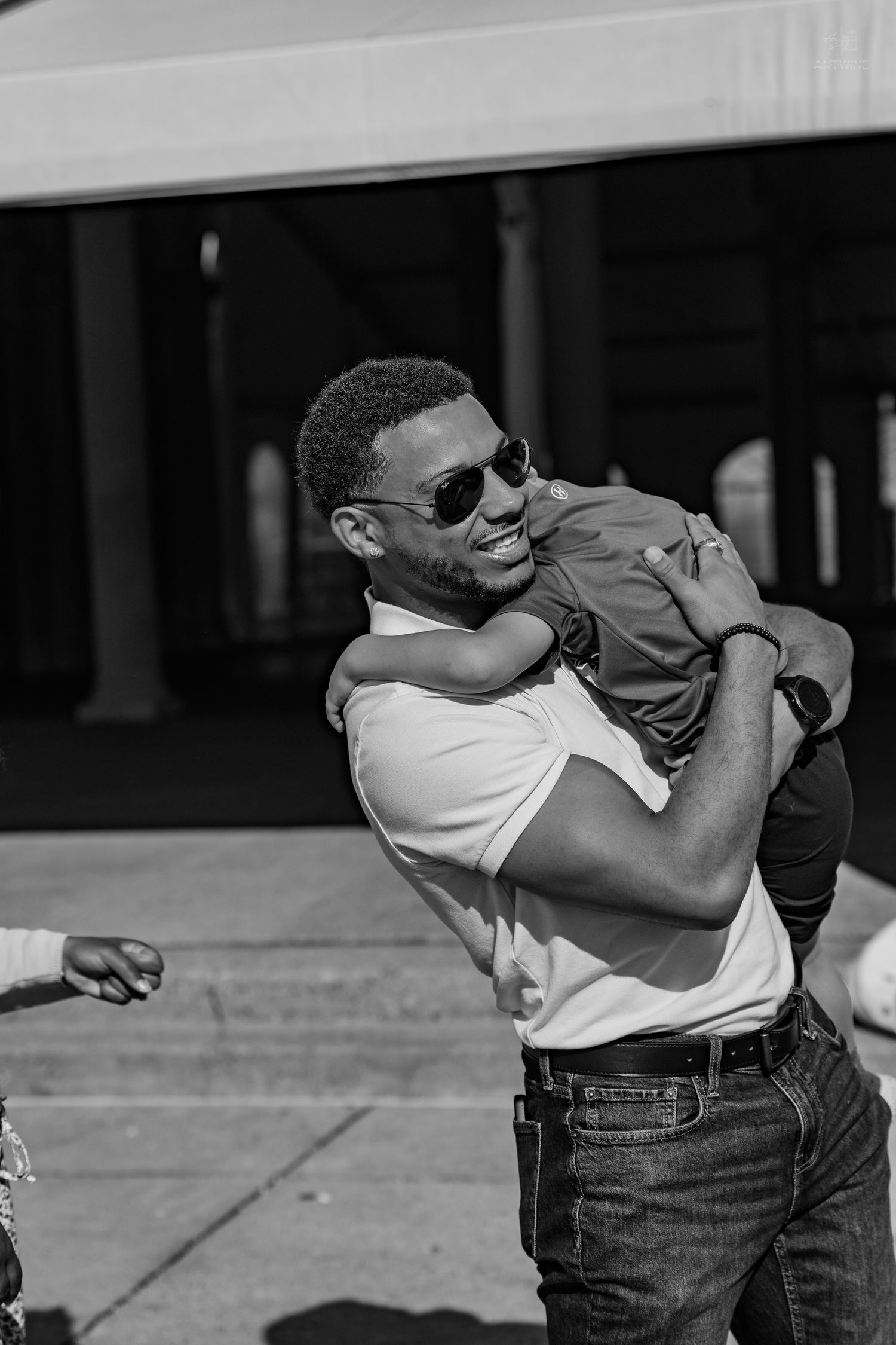 Black and white photo of a smiling man in sunglasses carrying a child over his shoulder outdoors.
