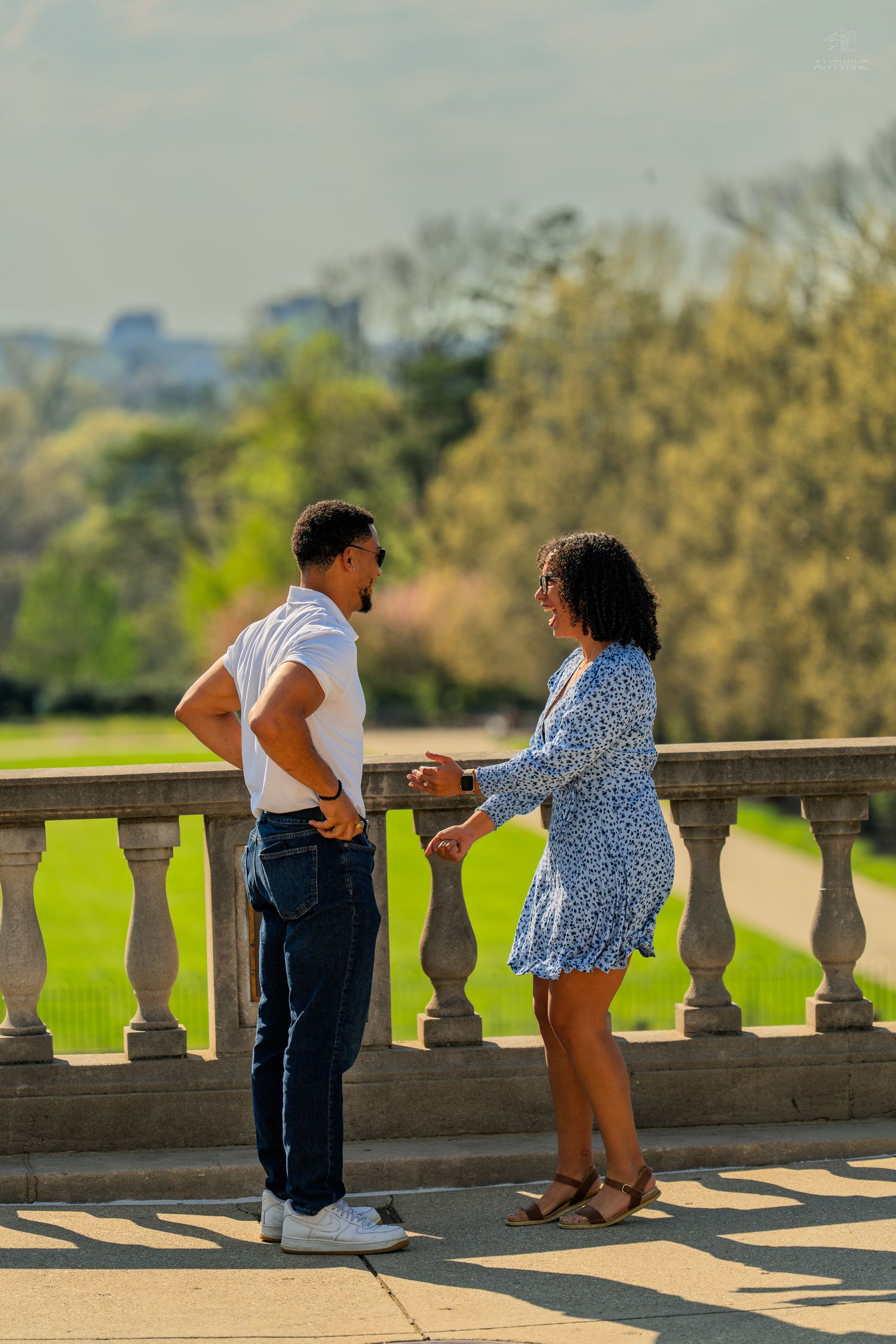 Couple holding hands and smiling together on a sunlit stone terrace with green park backdrop.