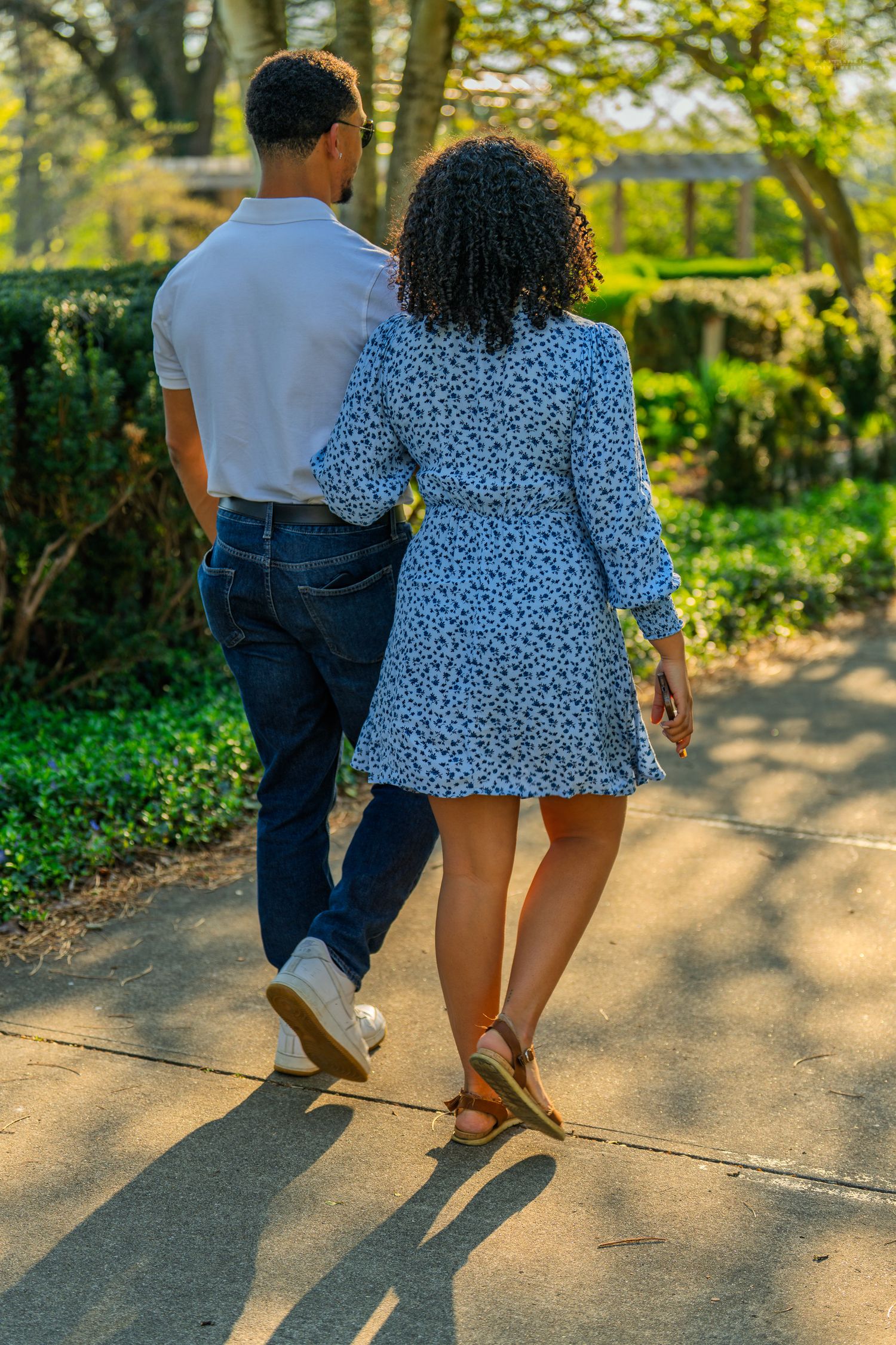 A couple strolling together down a sunlit park path, the woman in a floral dress holding his arm.