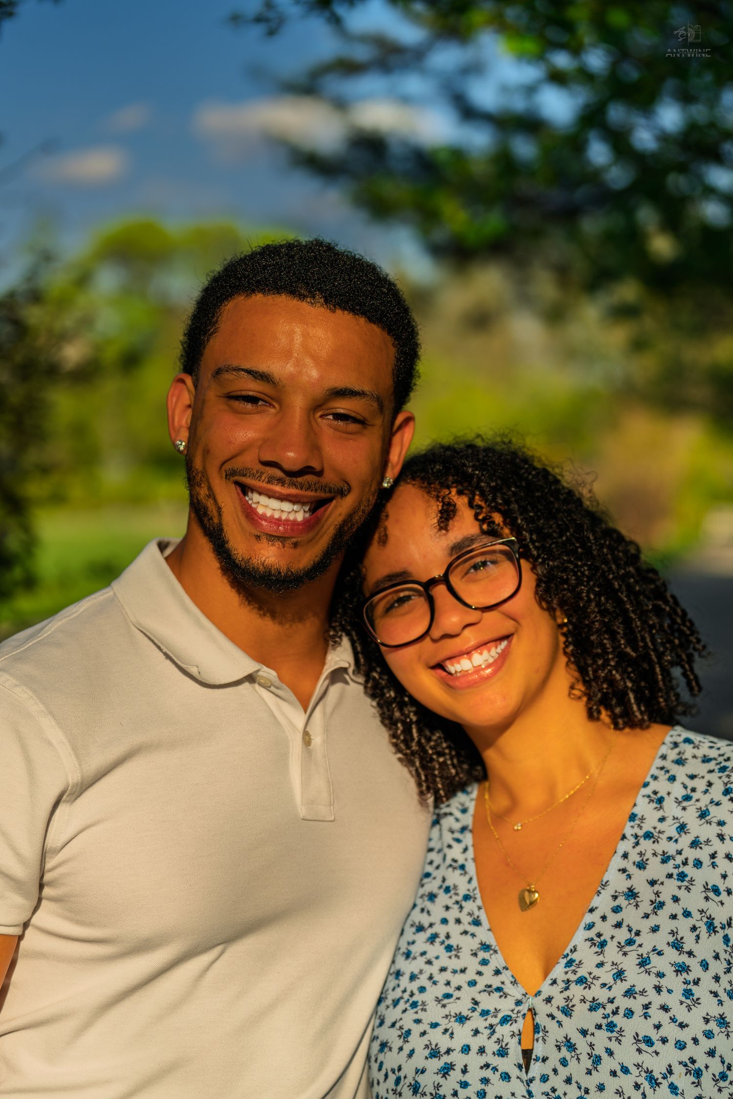 Happy couple posing cheek to cheek outdoors in golden sunlight, woman in blue floral dress smiling.
