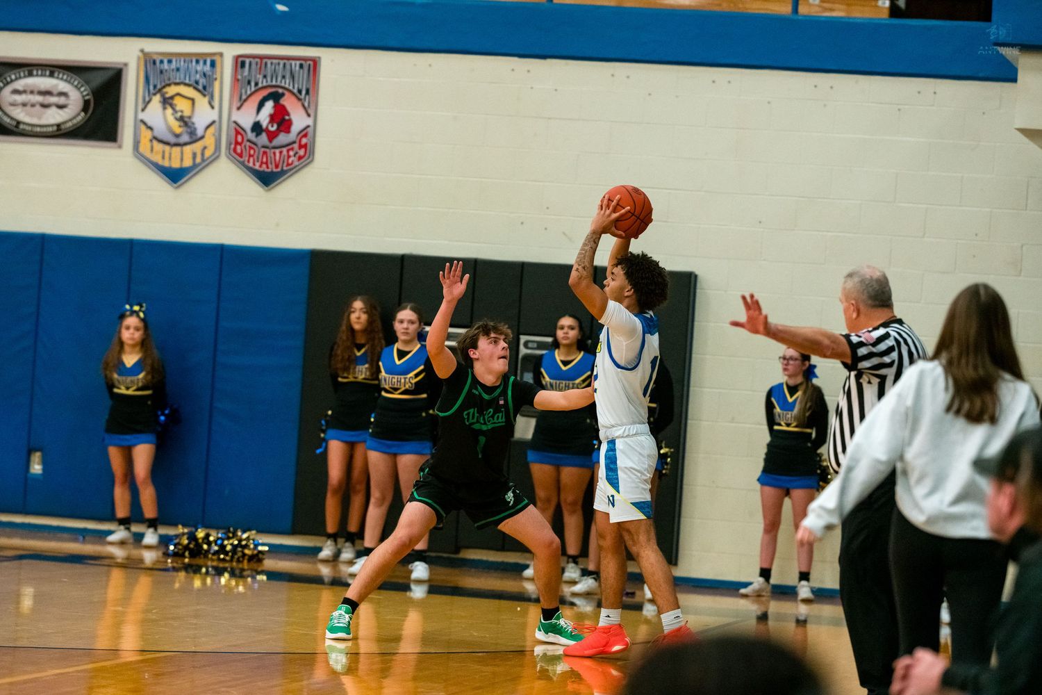 Basketball player shoots over defender while cheerleaders watch from sideline.