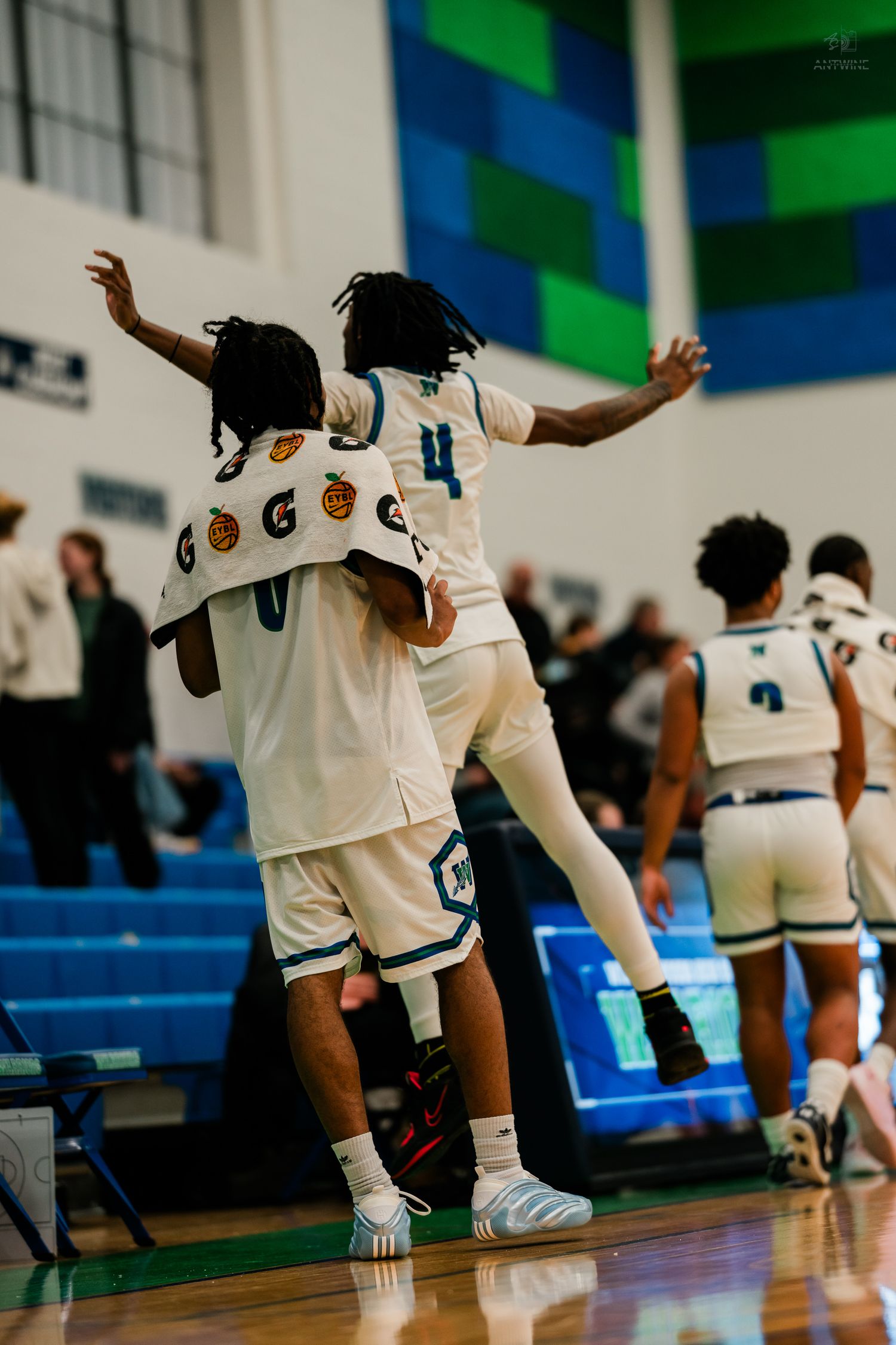 Two basketball players in white uniforms celebrate on the court, one jumping on teammates back with arms spread wide.