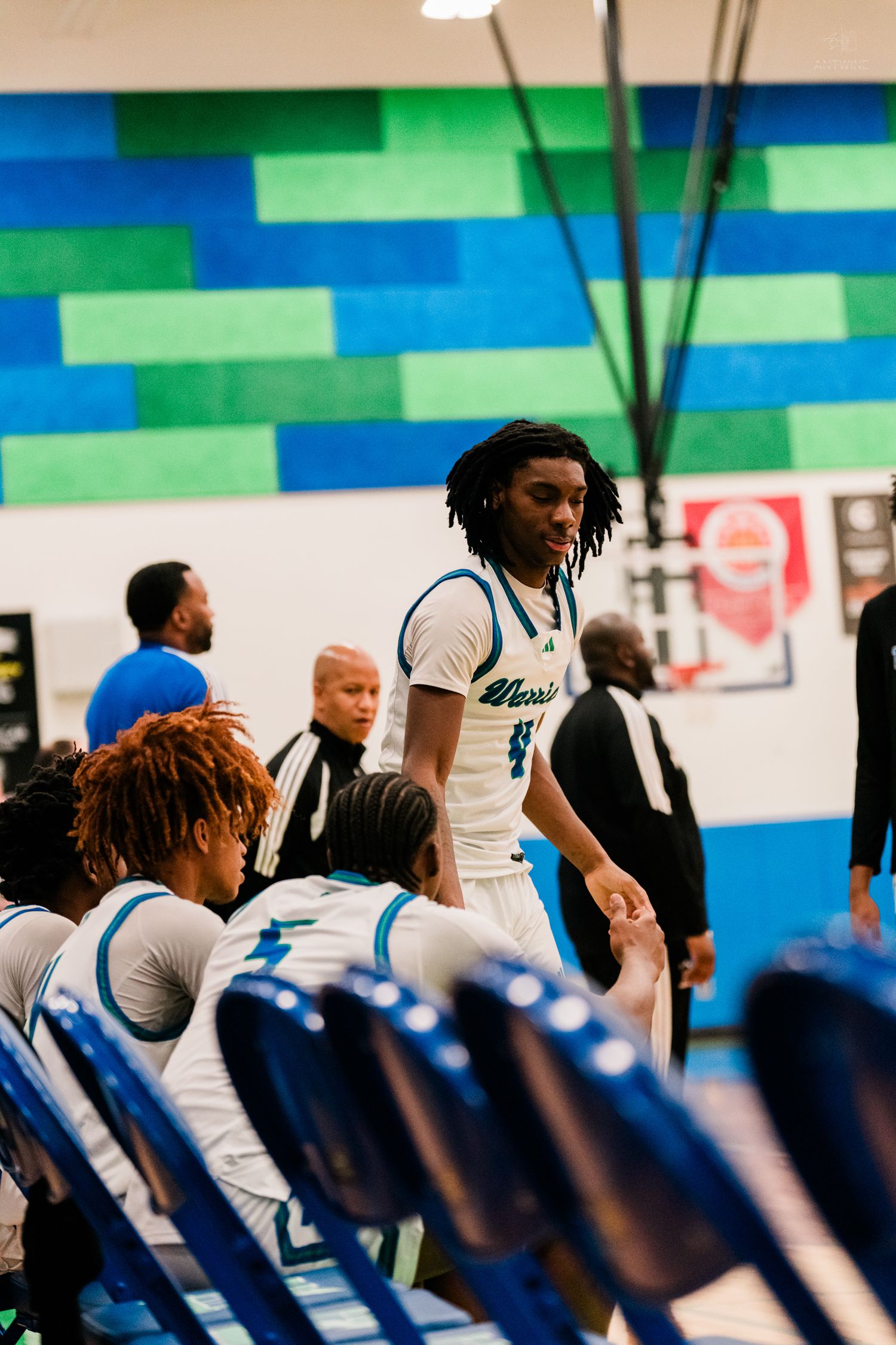 Tall basketball player with dreadlocks greets teammates near bench.